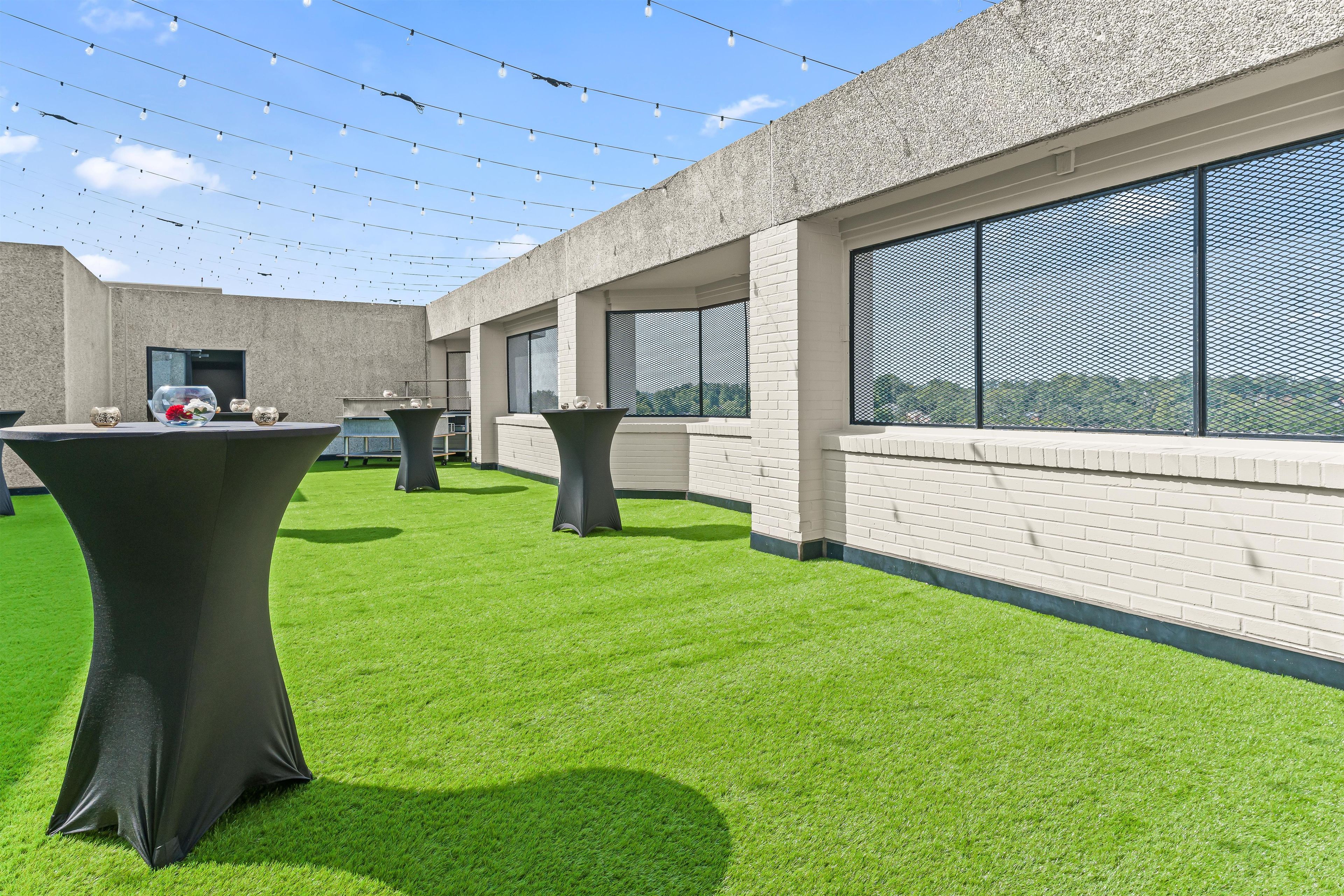 Un espacio al aire libre con césped artificial, mesas y sillas, y un edificio con ventanas grandes y una pared con textura.