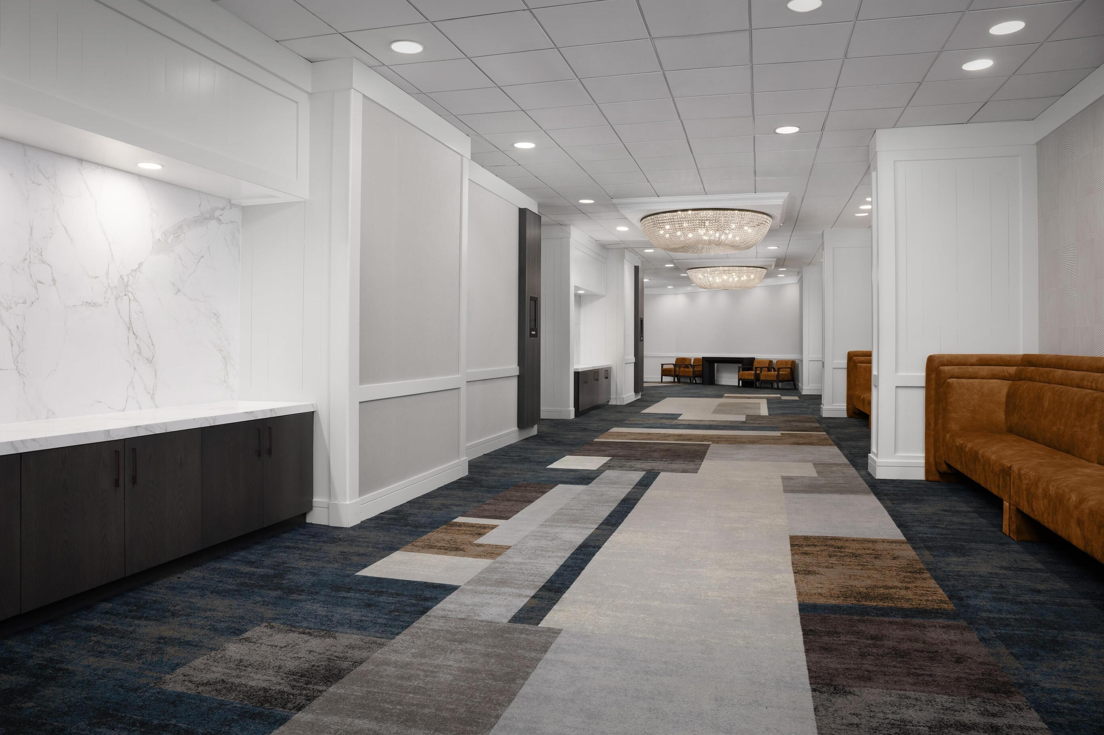 A modern hotel lobby with a marble reception desk, a patterned carpet, a chandelier, and a brown sofa.