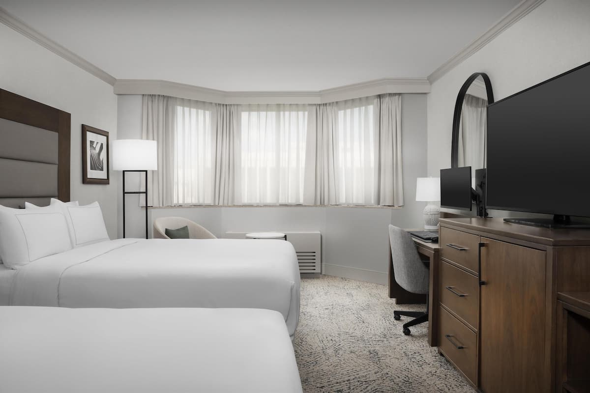 Habitación de hotel con dos camas, un escritorio, una silla, un televisor y un ventanal con cortinas.