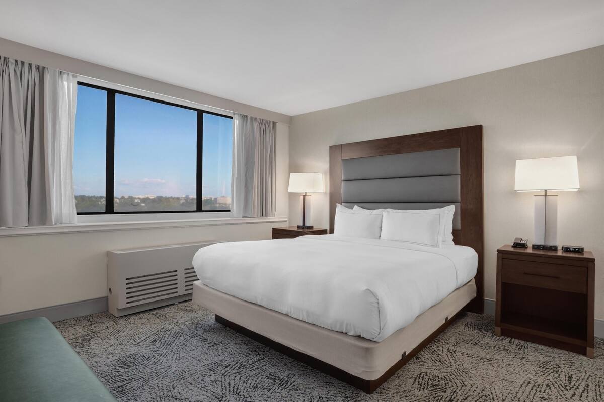 Habitación de hotel con una cama grande, dos mesitas de noche con lámparas, vista a la ciudad a través de un ventanal y un radiador.