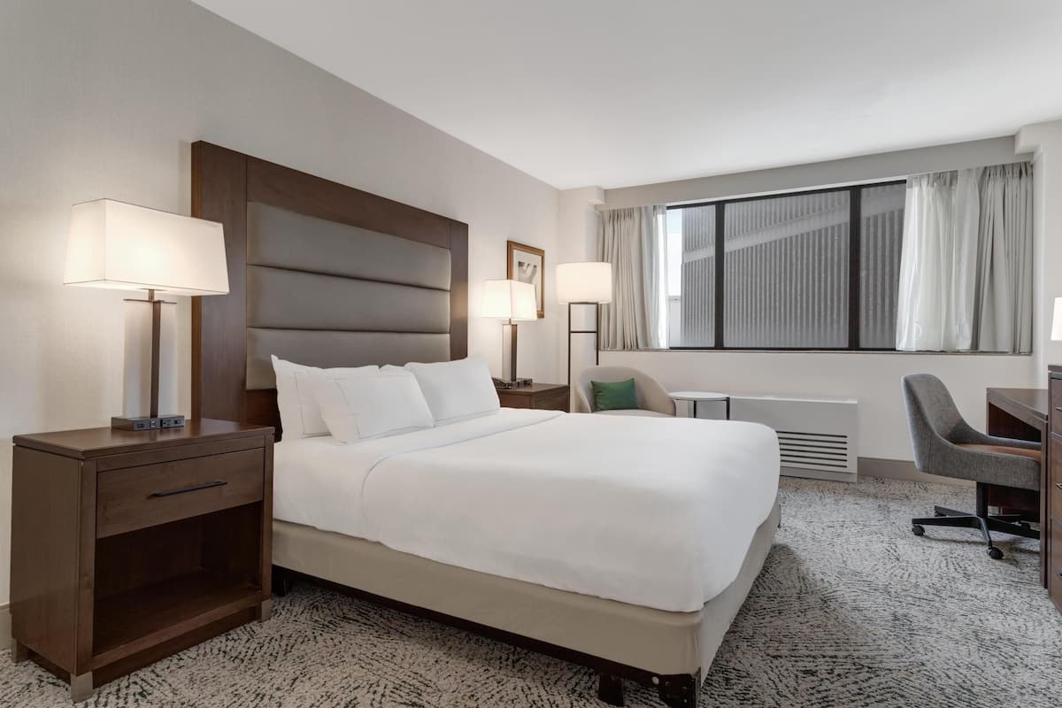 Una habitación de hotel con una cama grande, dos mesitas de noche con lámparas, un escritorio con una silla y un ventanal con cortinas.