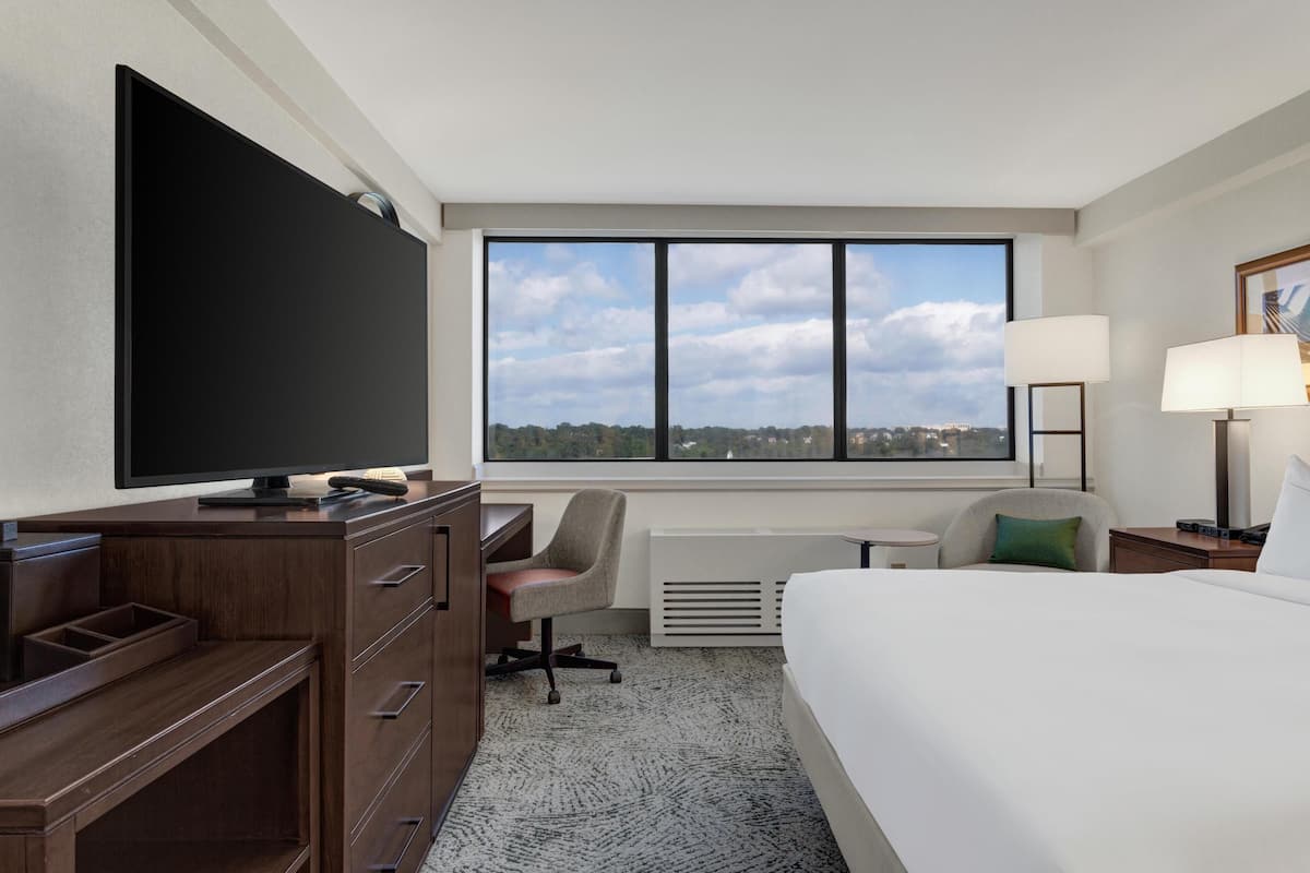 Habitación de hotel con un ventanal grande, un televisor de pantalla plana, un escritorio con silla, una cama y una mesita de noche con lámparas.
