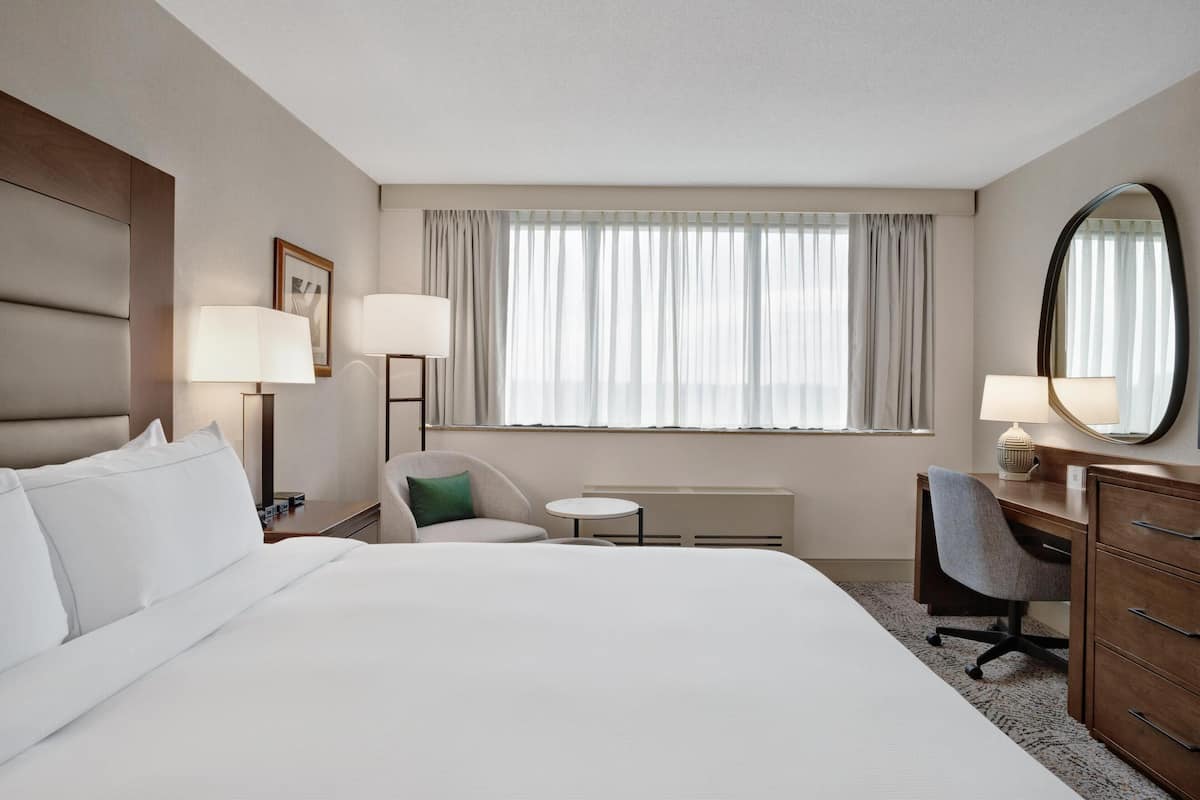 Habitación de hotel con una cama grande, un escritorio, una silla, una mesa redonda y un ventanal con cortinas.
