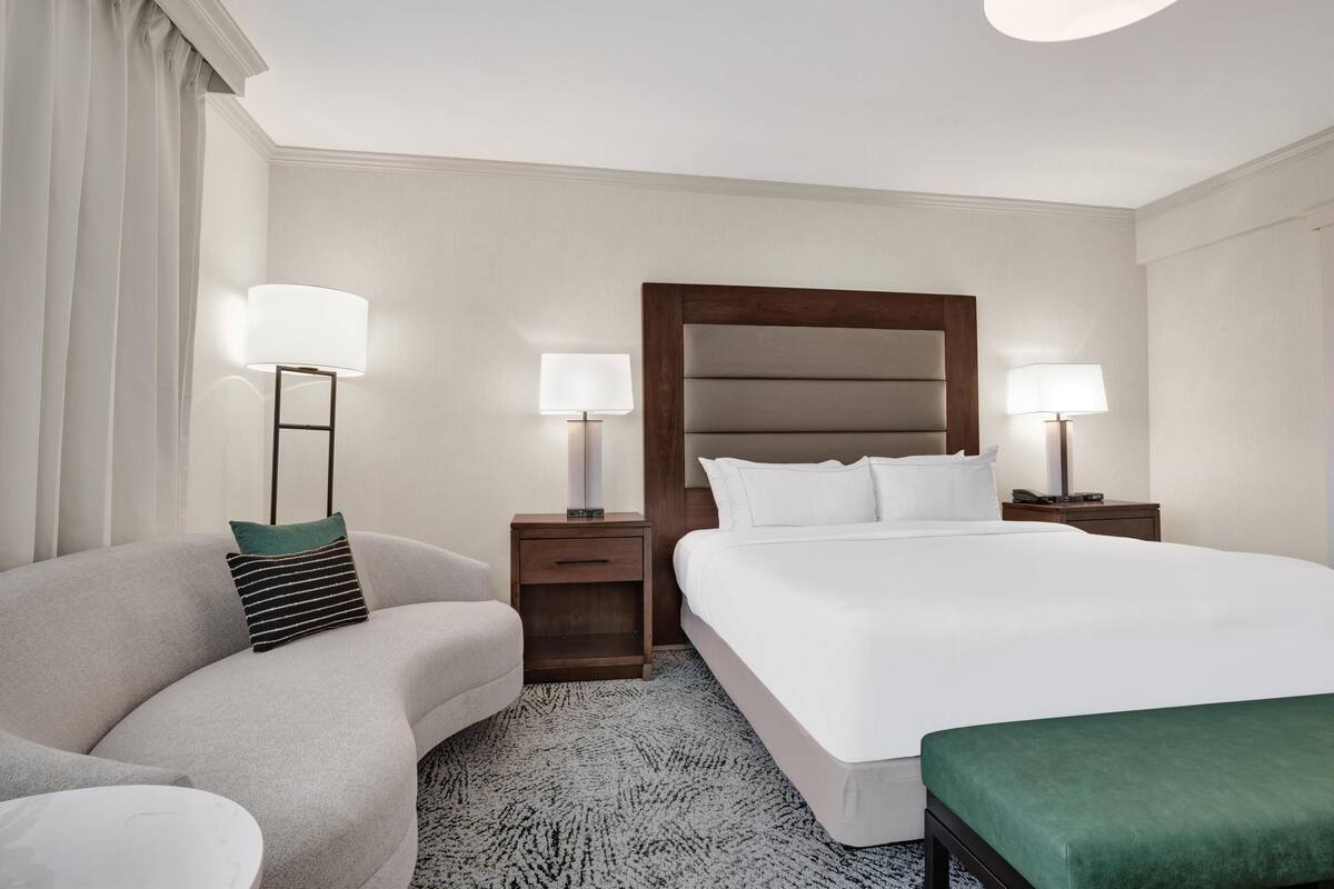 Habitación de hotel con una cama grande, un sofá, dos mesitas de noche con lámparas y piso alfombrado.