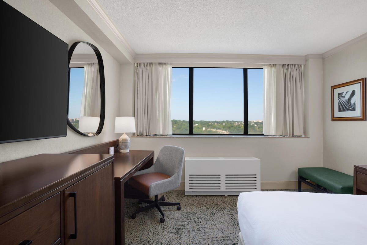 Habitación de hotel con una cama grande, un escritorio con silla, un espejo amplio, un radiador y una ventana con cortinas.