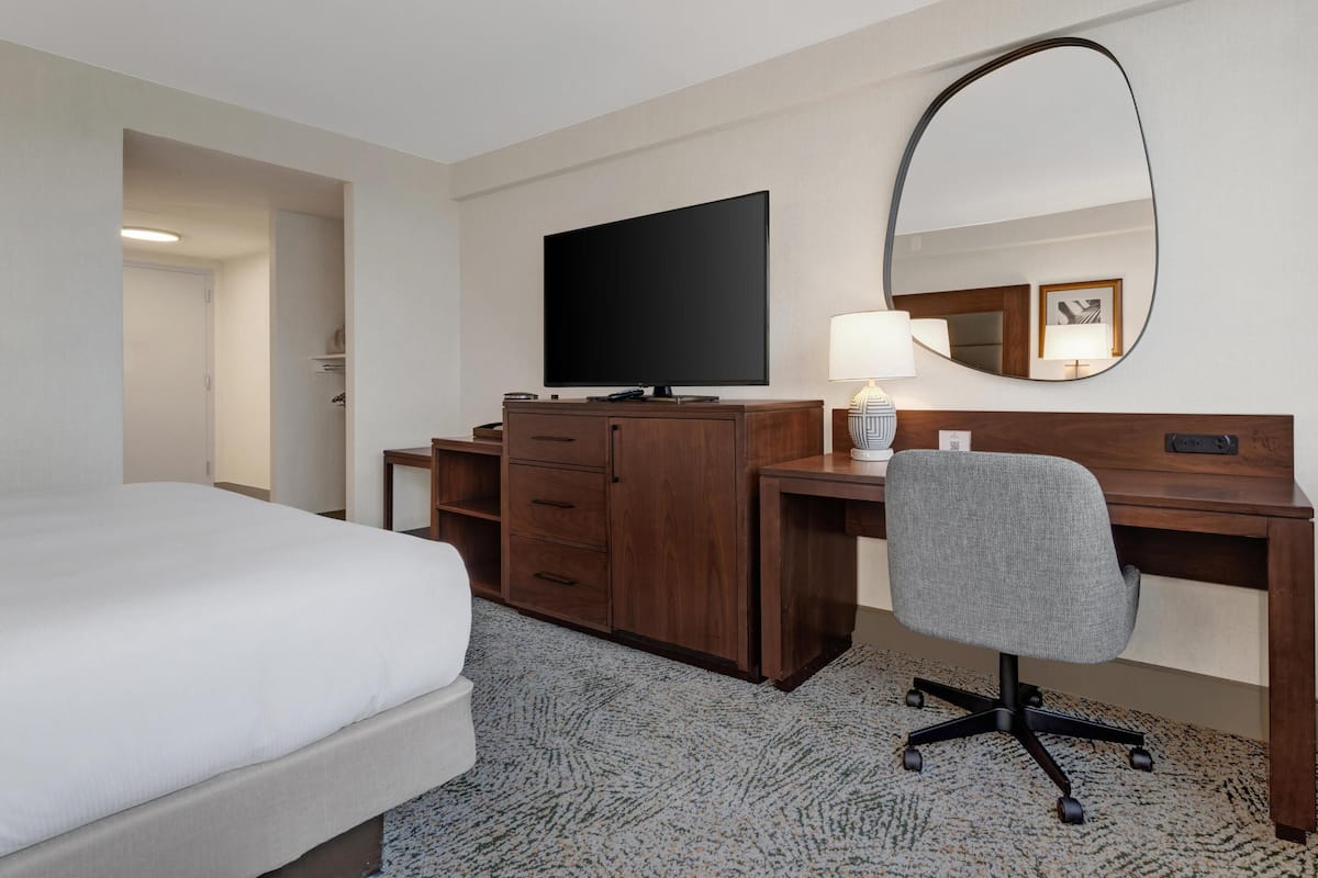 Habitación de hotel con cama, escritorio y silla, televisor, espejo y lámpara.