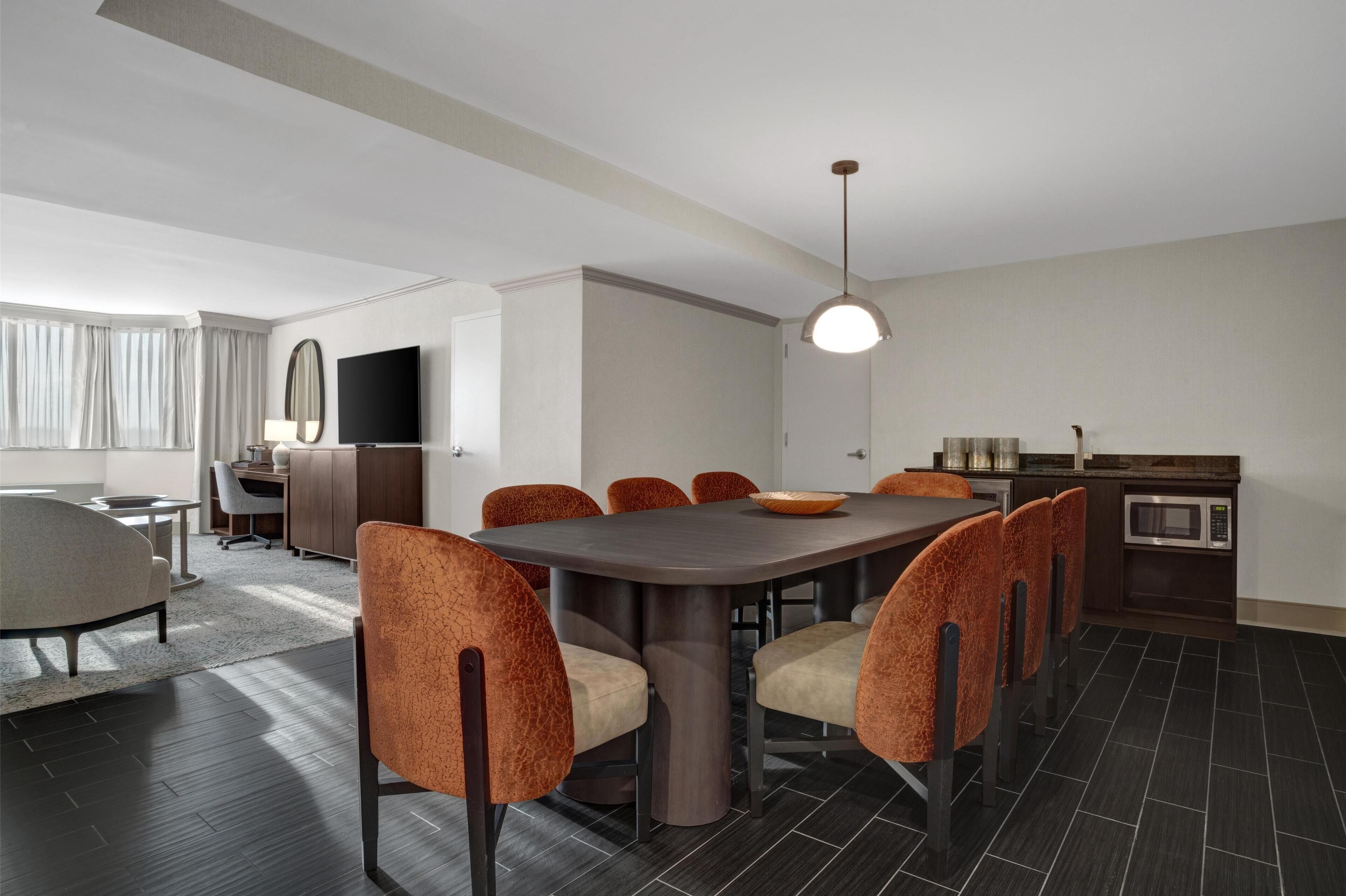 Una habitación de hotel moderna con comedor que incluye una mesa de madera oscura y sillas tapizadas en naranja, una cocina con microondas y un gran ventanal con cortinas.