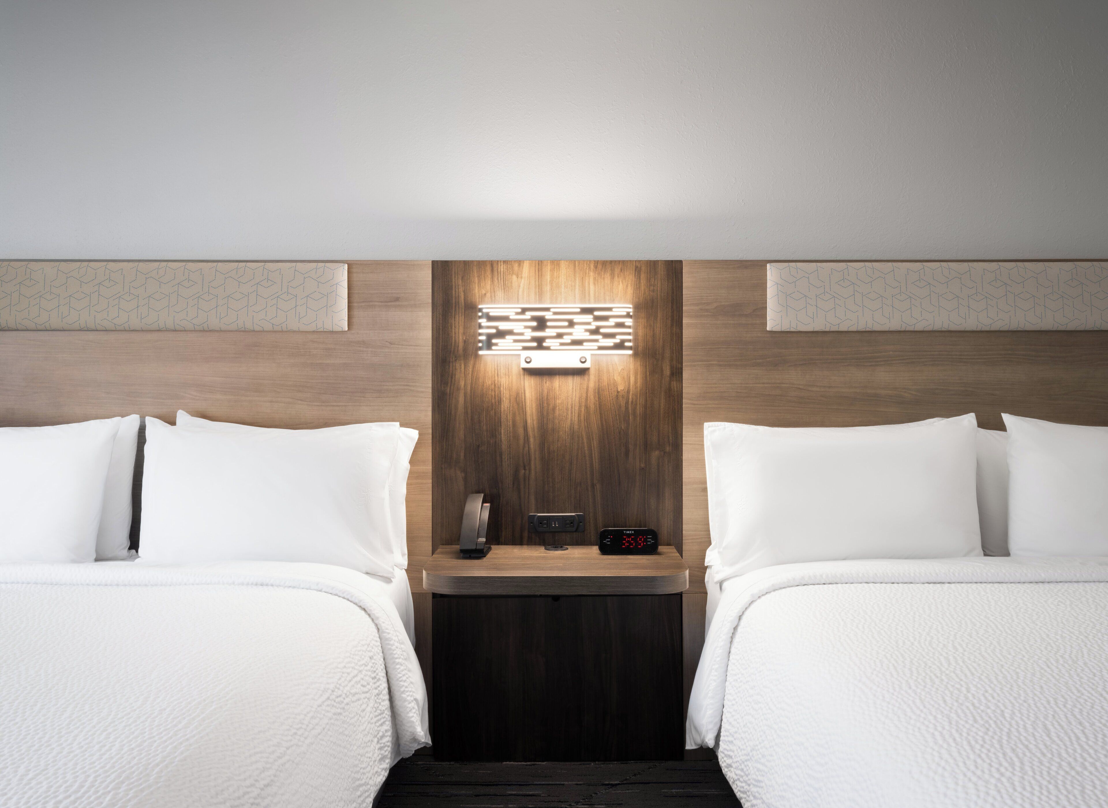 Habitación de hotel con dos camas, una mesita de noche con un reloj y una lámpara fijada en la pared.