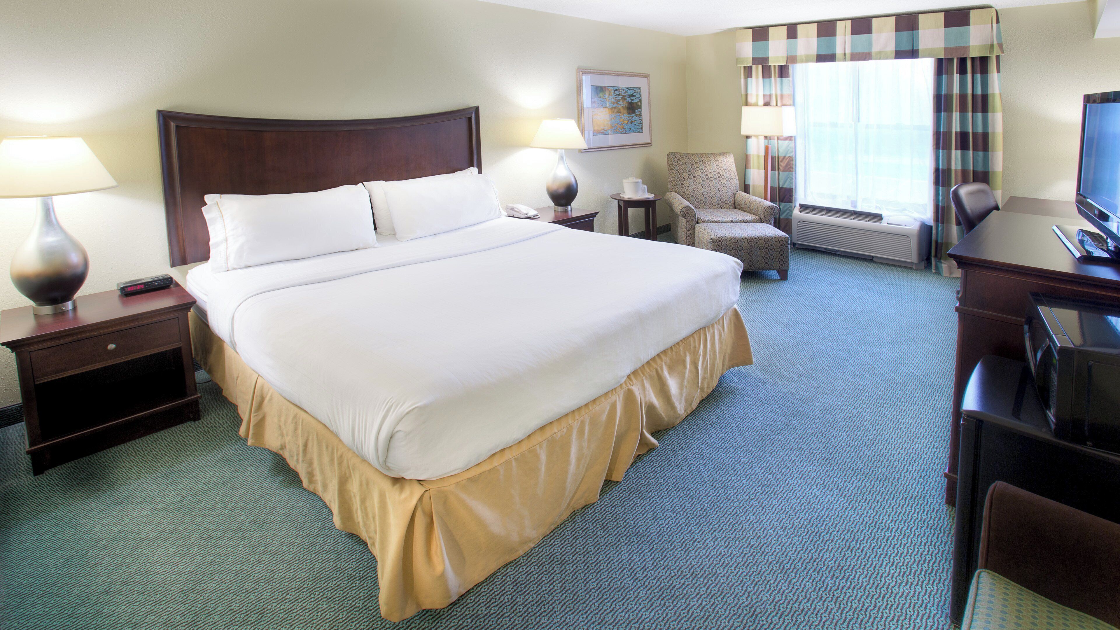 Ein Hotelzimmer mit einem großen Bett, einem Schreibtisch mit Computer, einem Sessel, einer Lampe und einem Gemälde an der Wand.