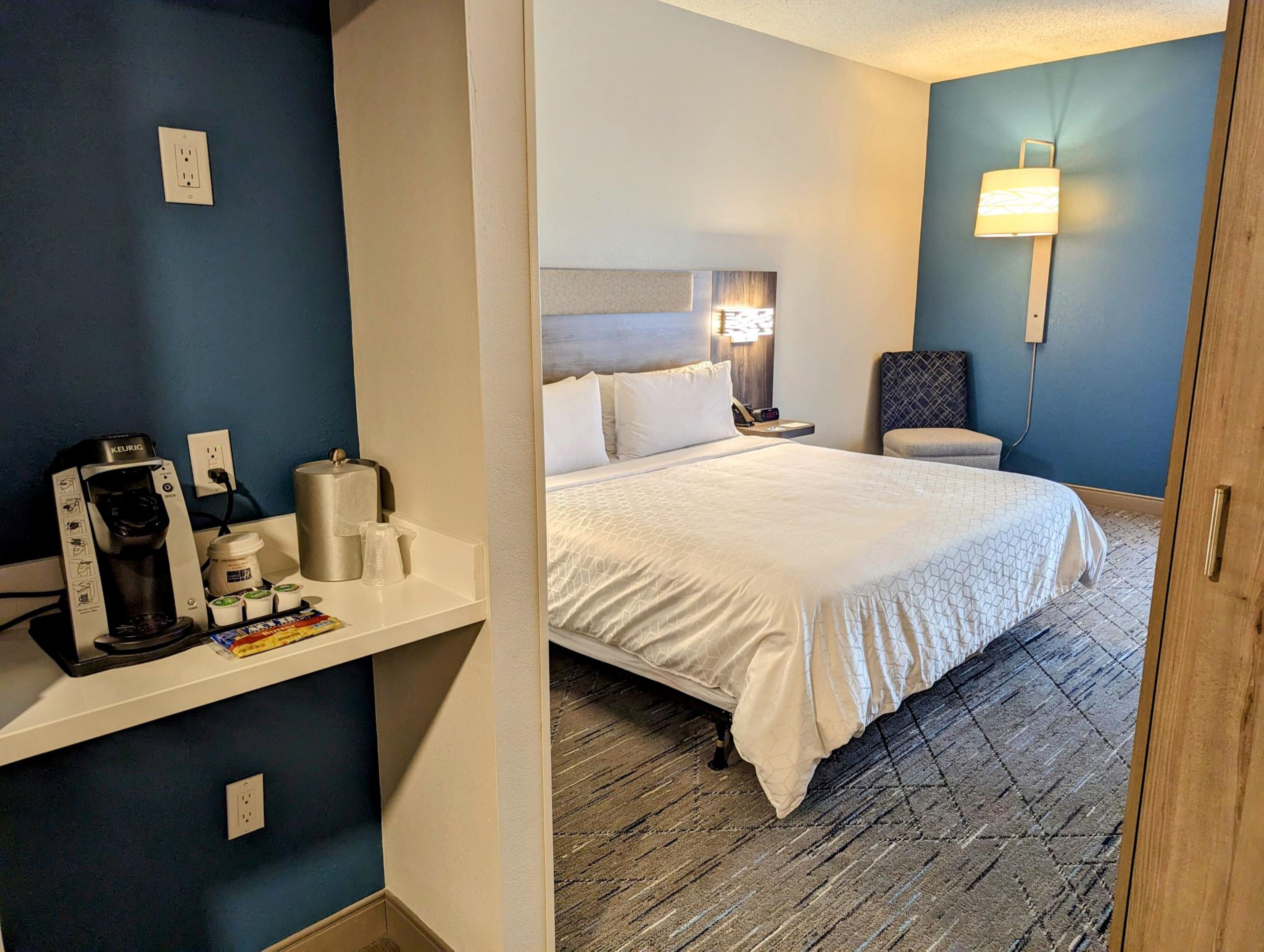 A hotel room with a bed, a nightstand, a chair, a coffee maker, and a lamp.