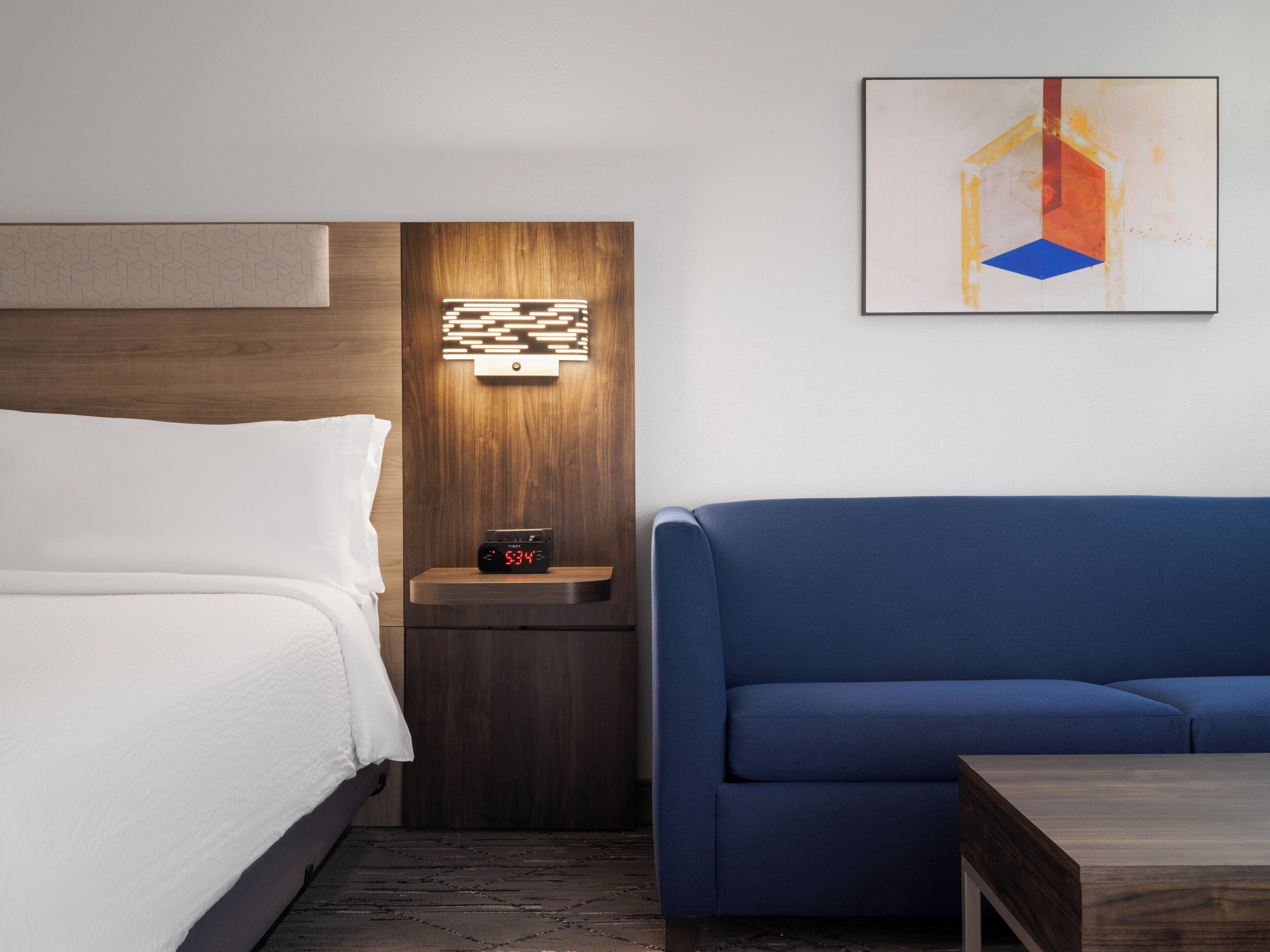 A hotel room with a bed, a nightstand with a clock, a blue sofa, and a wooden headboard.