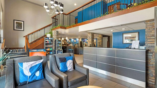 Un moderno vestíbulo de hotel con mostrador de recepción, zona de estar, escalera y vistas al área de la cocina.