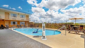 Un área de piscina con sillas de descanso, un trampolín y un edificio con múltiples ventanas y puertas.