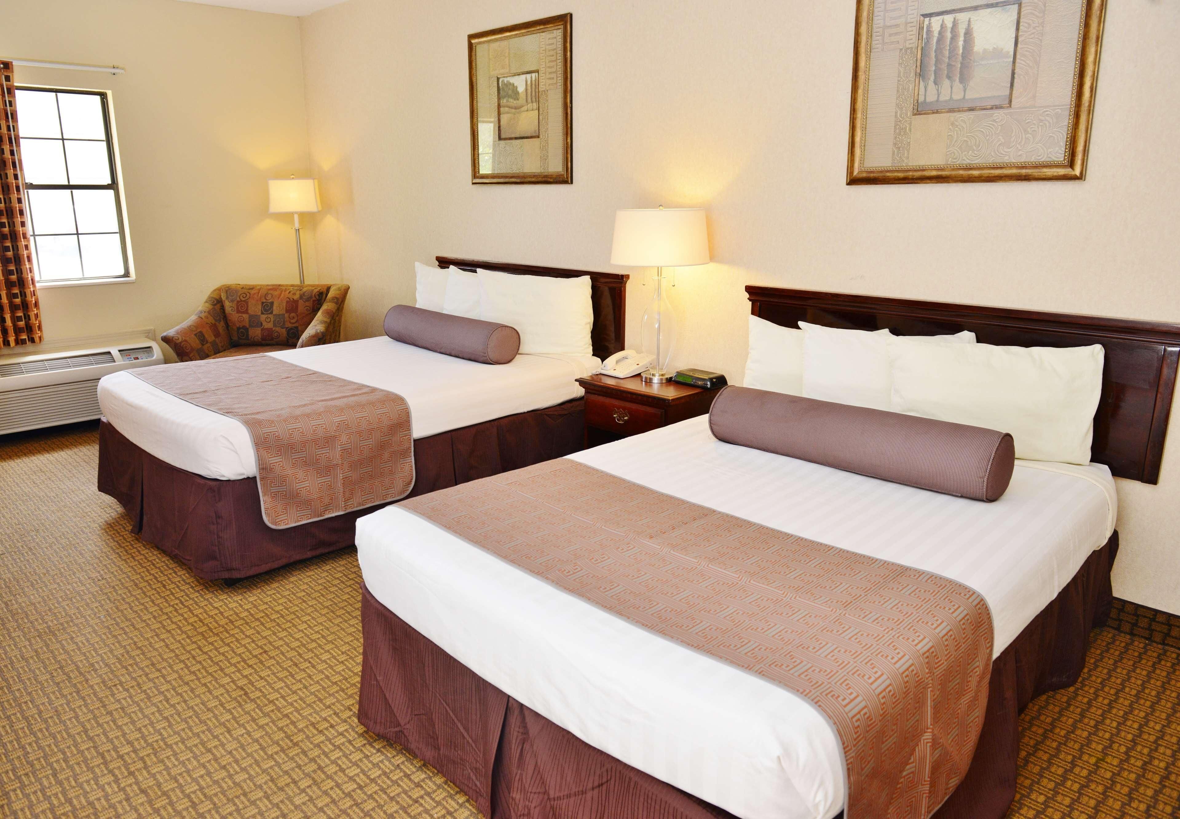 A hotel room with two beds, a chair, a nightstand with a lamp, and a framed picture on the wall.
