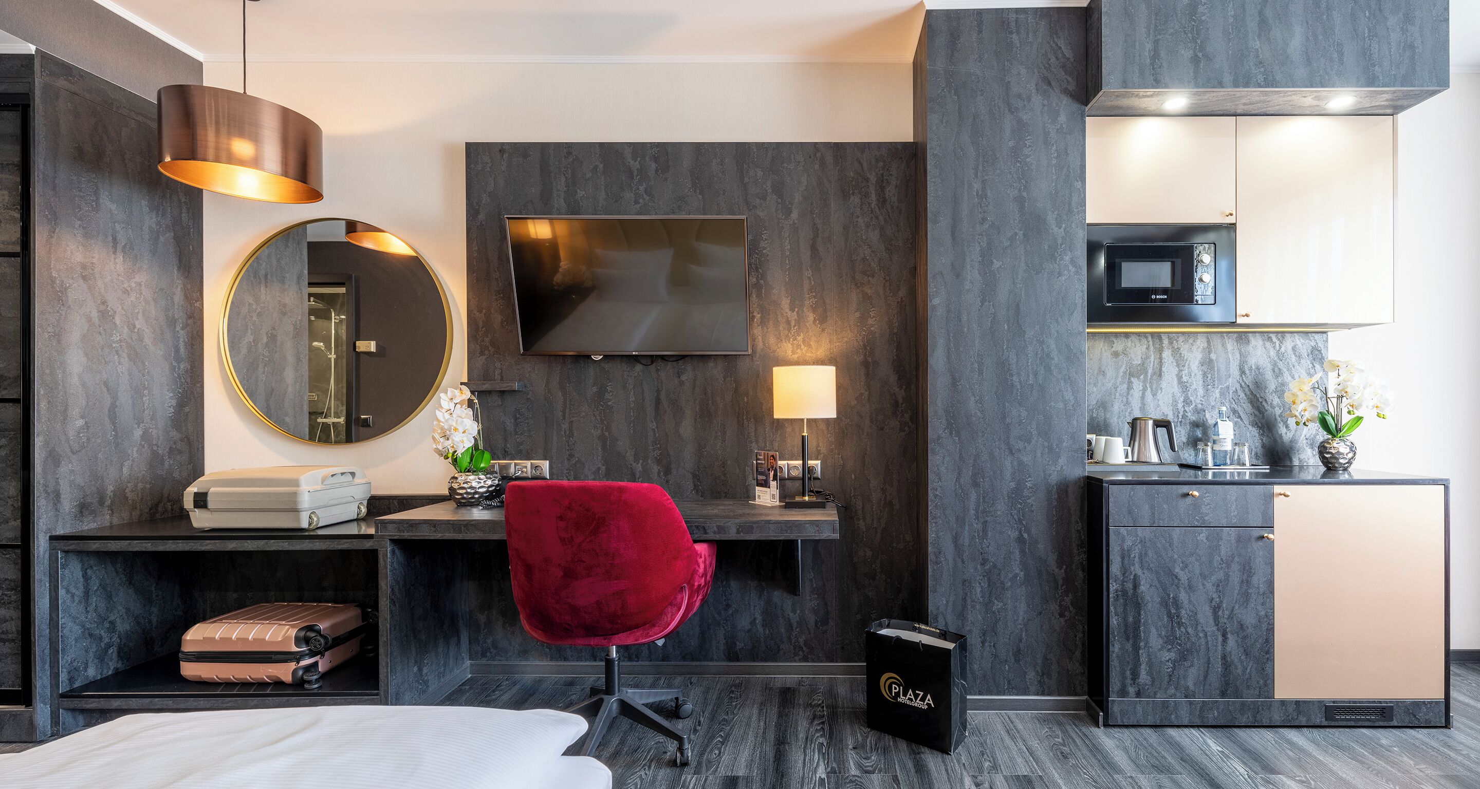 A modern hotel room with a bed, a desk with a computer, a TV, a mirror, a red chair, and a minibar.