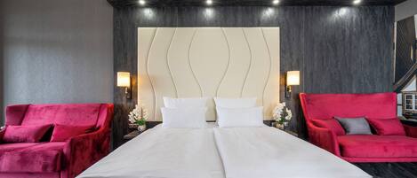 A hotel room with a large bed, two red armchairs, a modern headboard, and wall-mounted lamps.