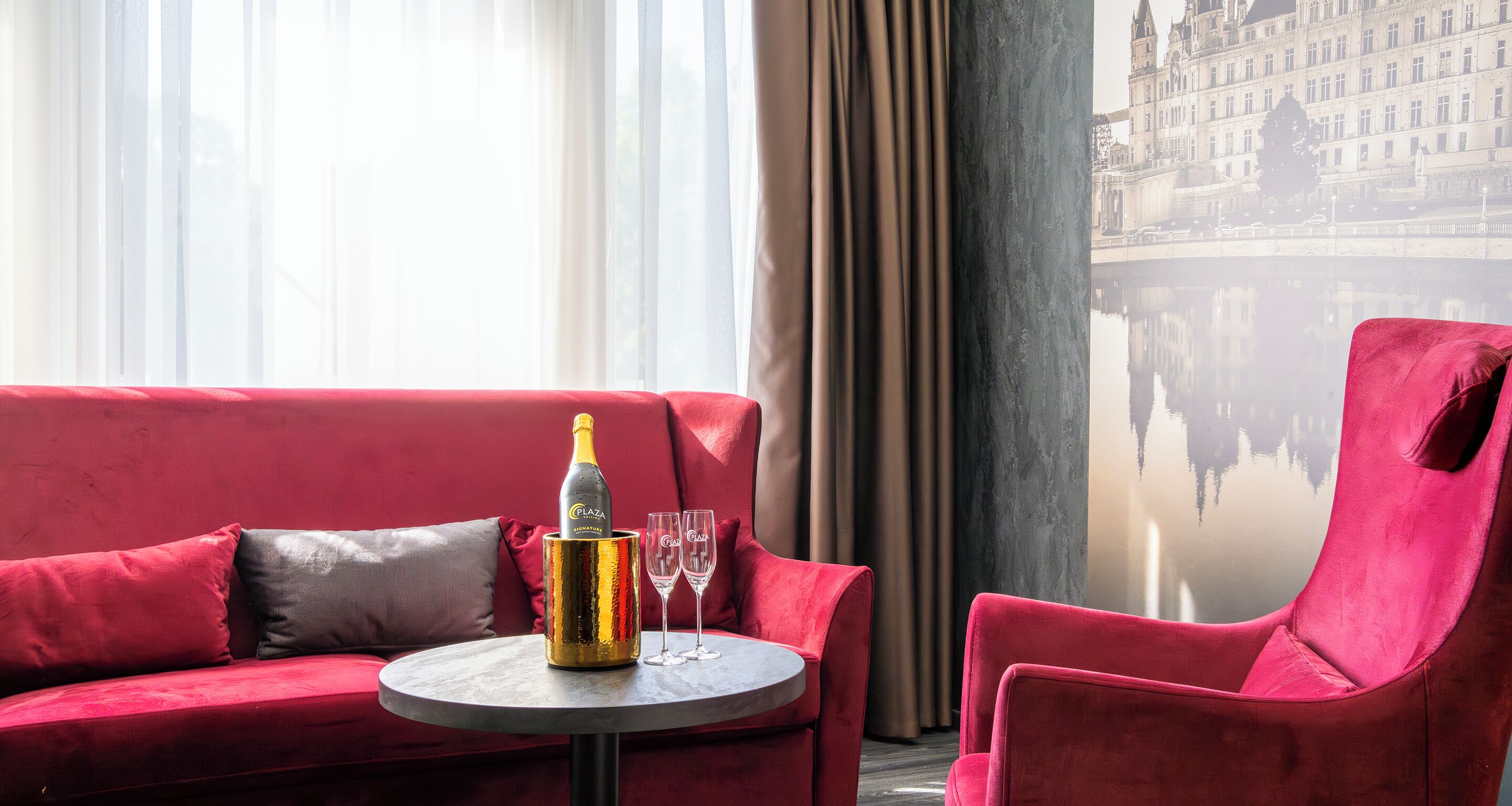 Ein rotes Sofa mit einer goldfarbenen Flasche und zwei Gläsern auf einem kleinen runden Tisch, mit Blick auf ein Gebäude durch ein Fenster.