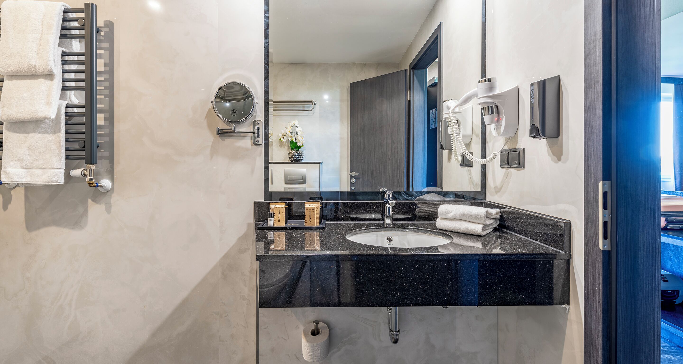 A modern bathroom with a black granite countertop, a round mirror, a towel rack, a hairdryer, and a toilet paper holder.