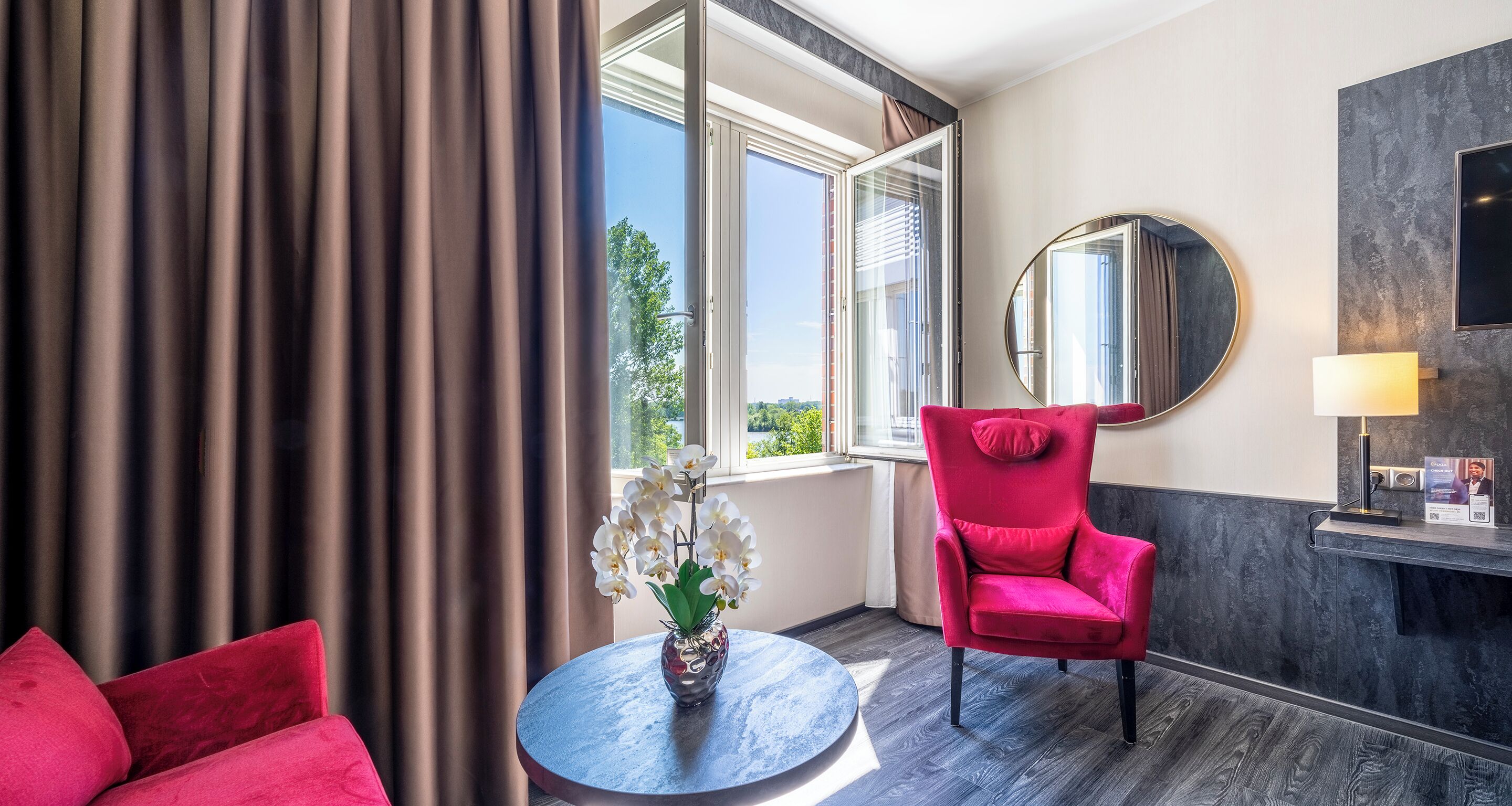 A hotel room with a red chair, a round table with a vase of flowers, a large mirror, and a view of the outdoors through a window.