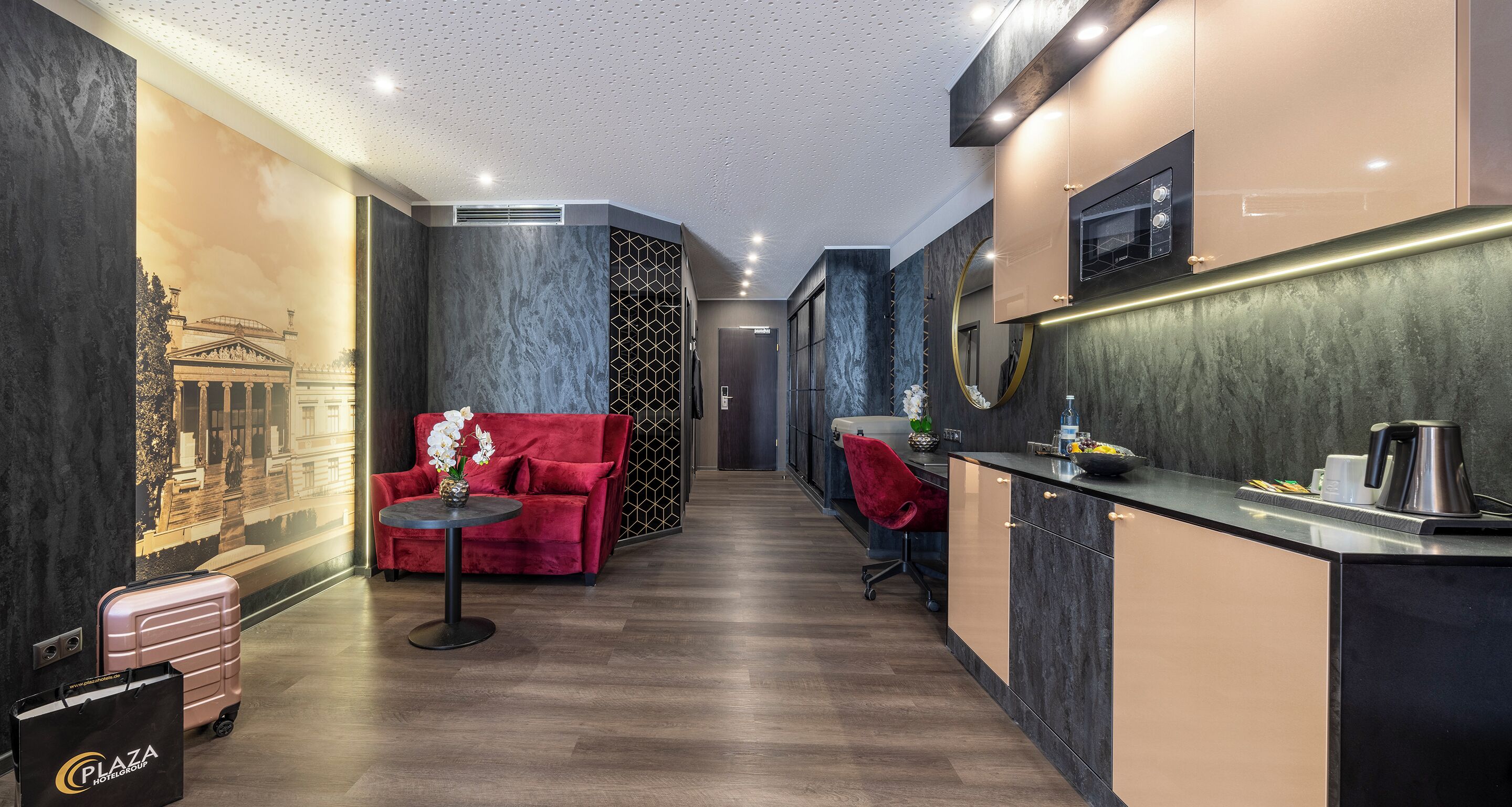 A modern hotel corridor with a red sofa, a small table with flowers, a kitchenette, and a large mural on the wall.