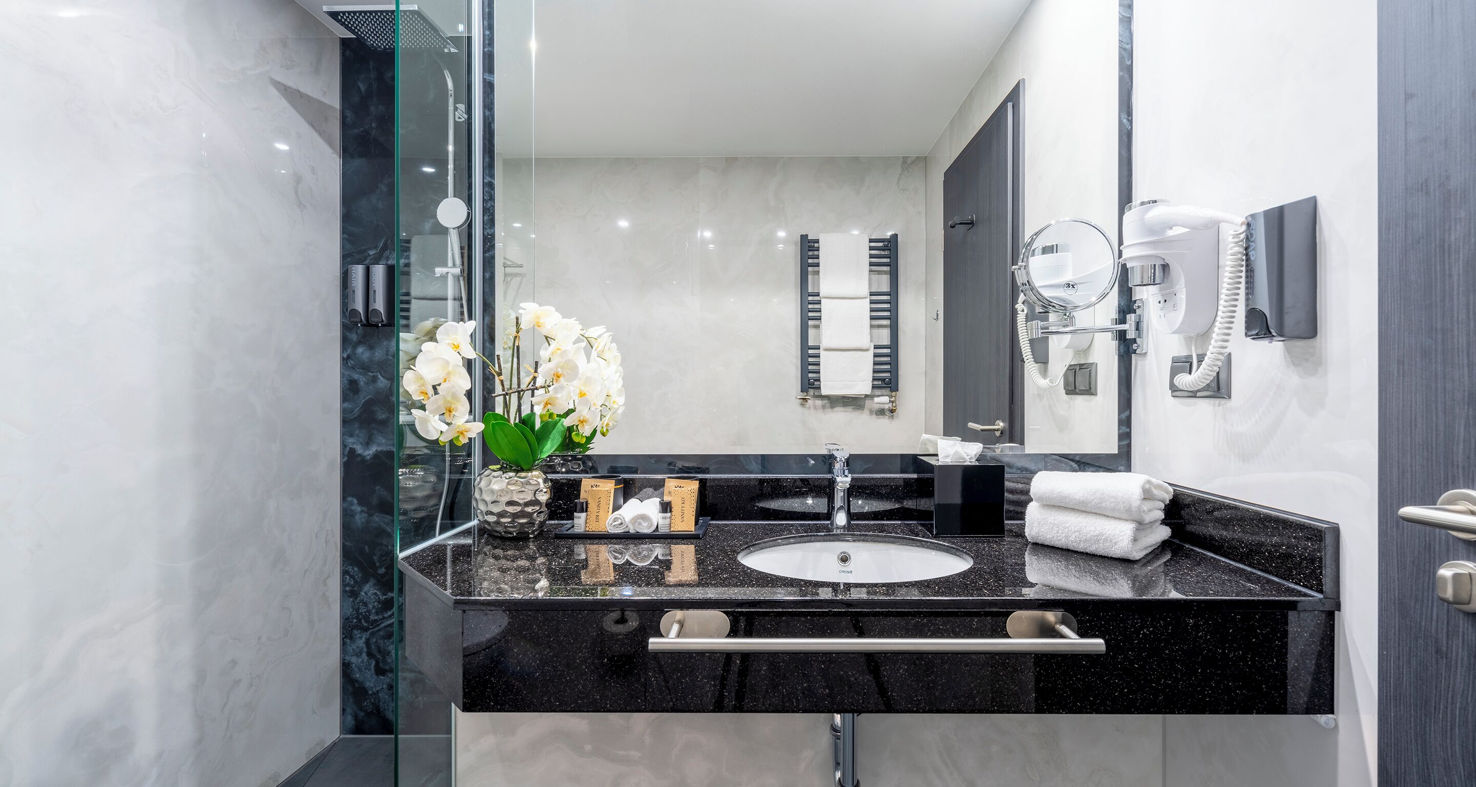 Un baño moderno con un lavabo de granito negro, un espejo grande, un cabezal de ducha de mano y un perchero para toallas.