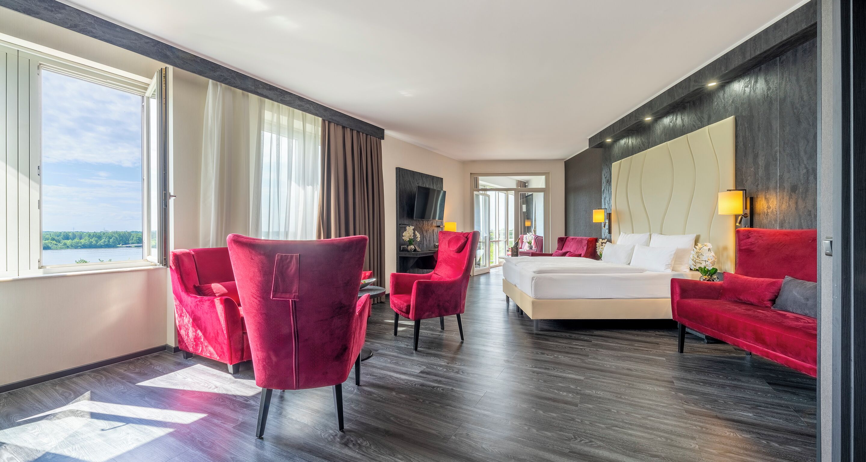 Een moderne hotelkamer met een groot bed, rode fauteuils, een rode bank, een televisie en uitzicht op de omgeving.