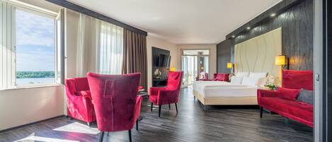 Een moderne hotelkamer met een groot bed, rode fauteuils, een rode bank, een televisie en uitzicht op de omgeving.