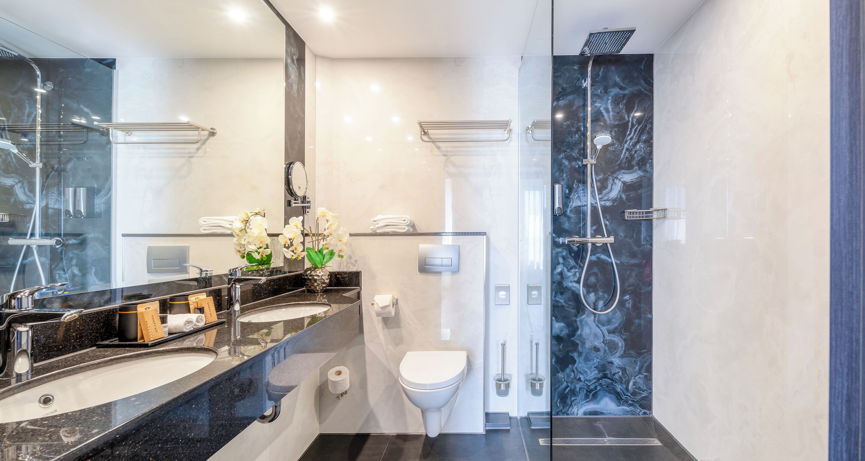 A modern bathroom with dual sinks, a toilet, a shower, and a large mirror.