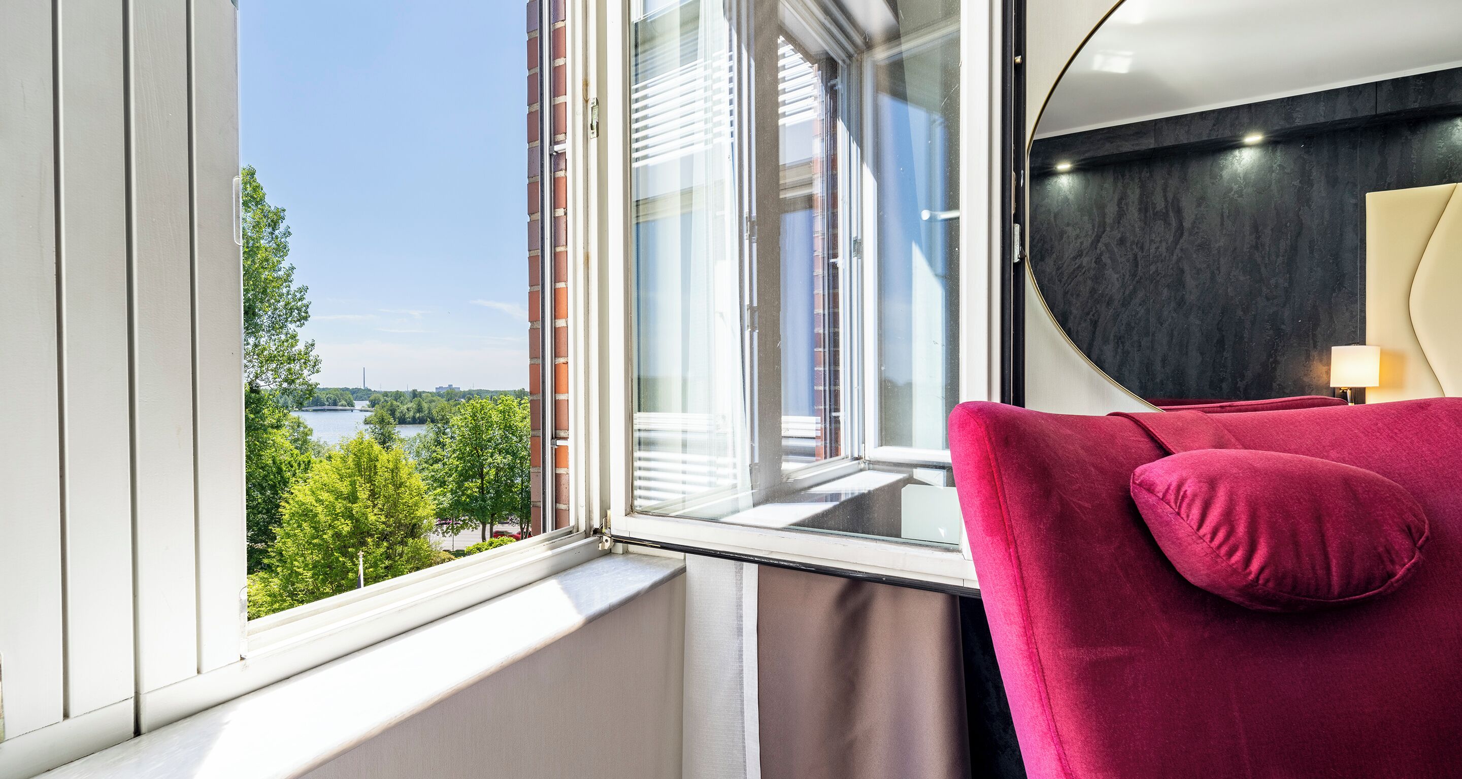 A room with a view of a river and trees, a window with blinds, a red chair, and a lamp.