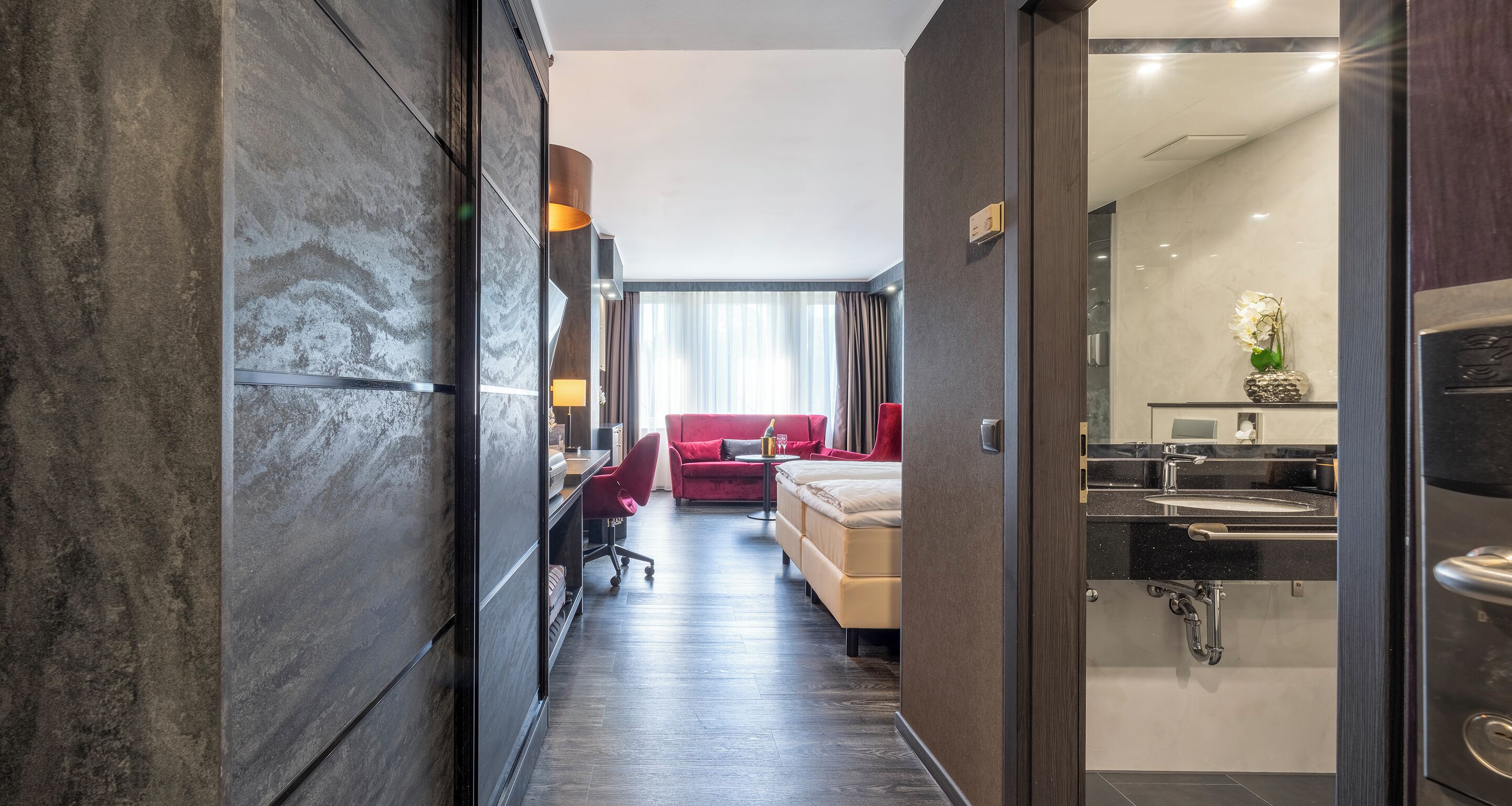Een moderne hotelkamer met een donker houten vloer, een rode bank, een bureau met stoel, en een groot raam met gordijnen.