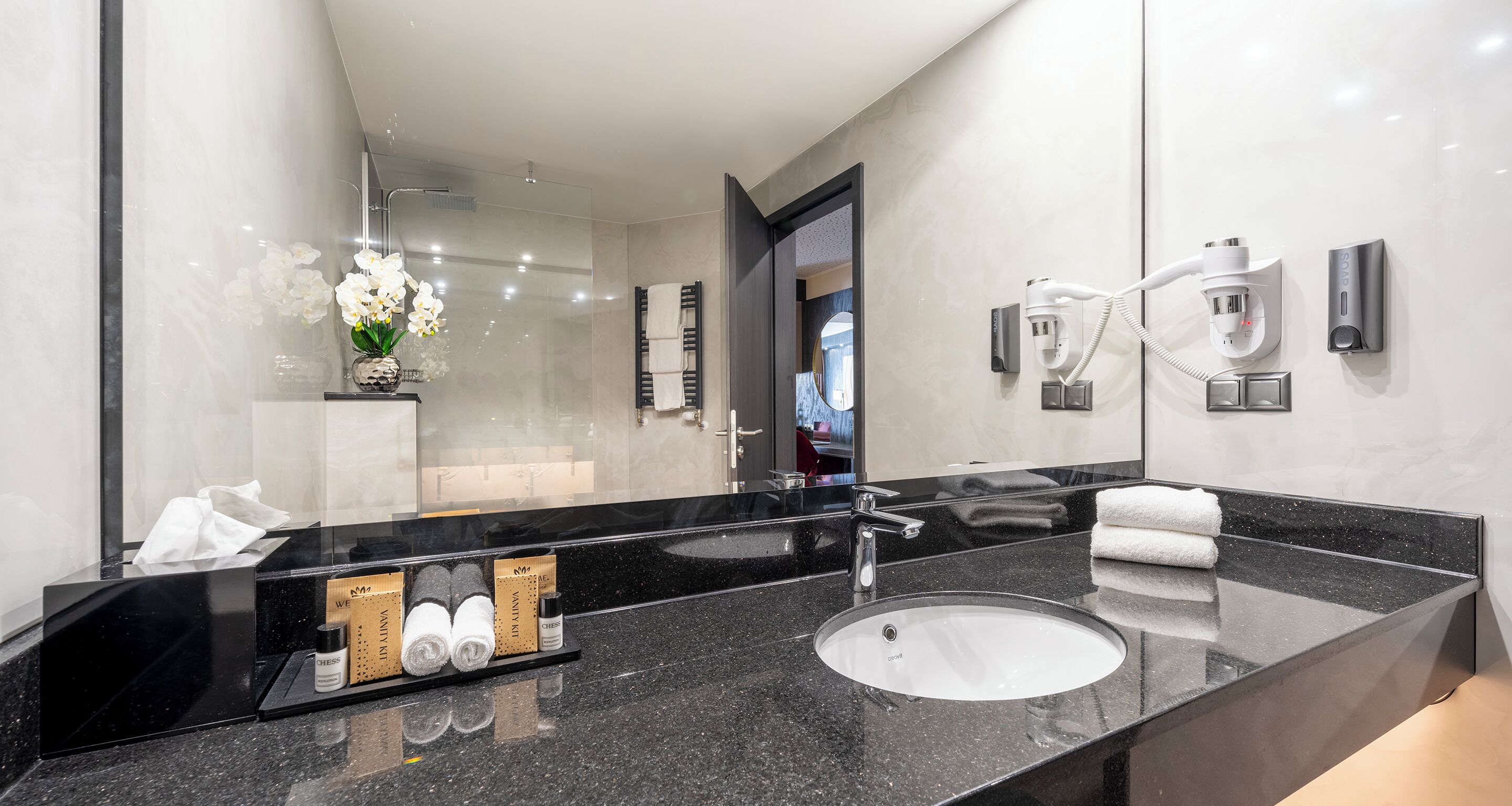 A modern bathroom with a black granite countertop, a white sink, and a large mirror. There are towels and toiletries on the counter.