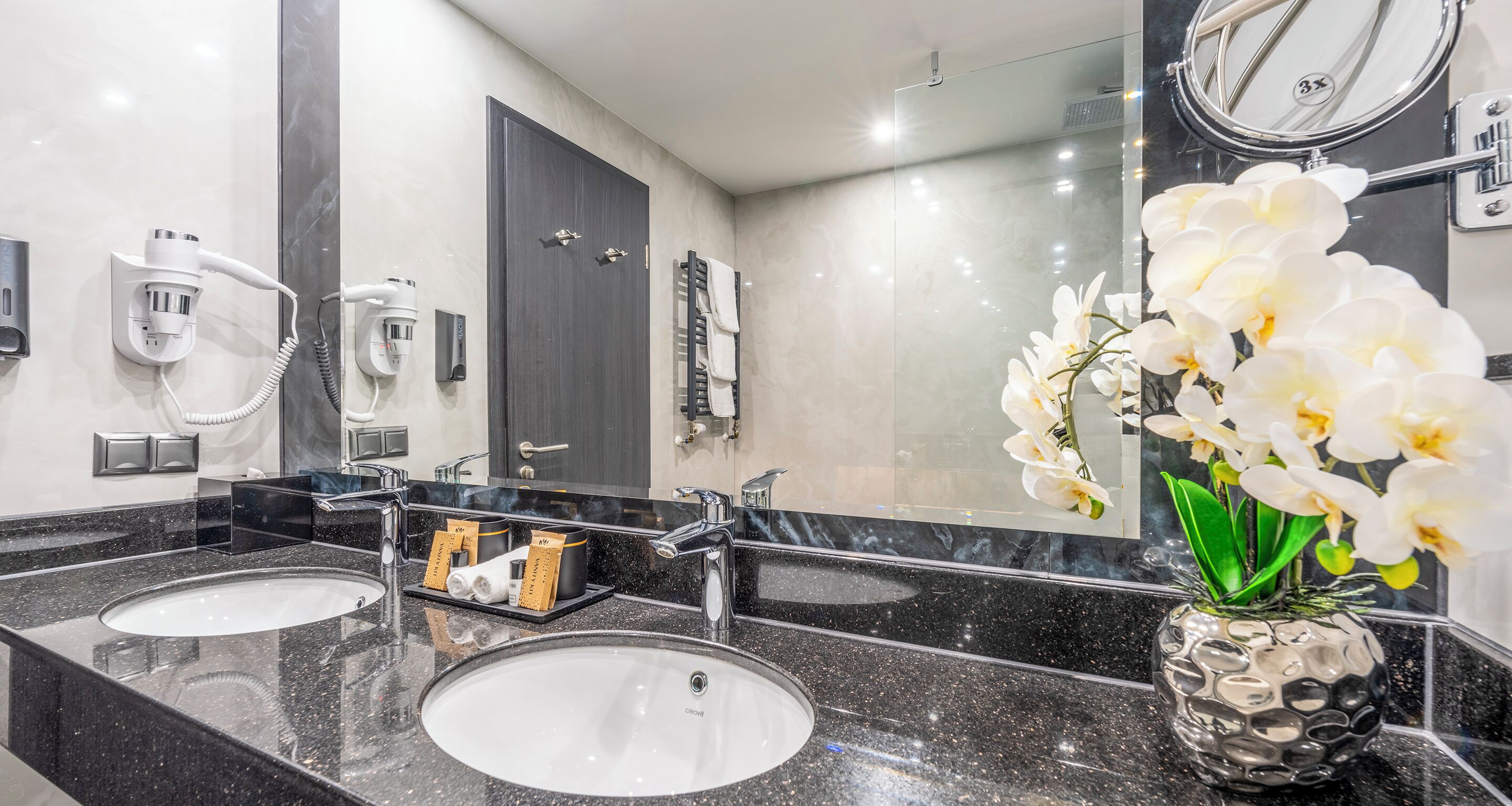 Un baño moderno con dos lavamanos, un espejo grande y un florero con flores blancas.