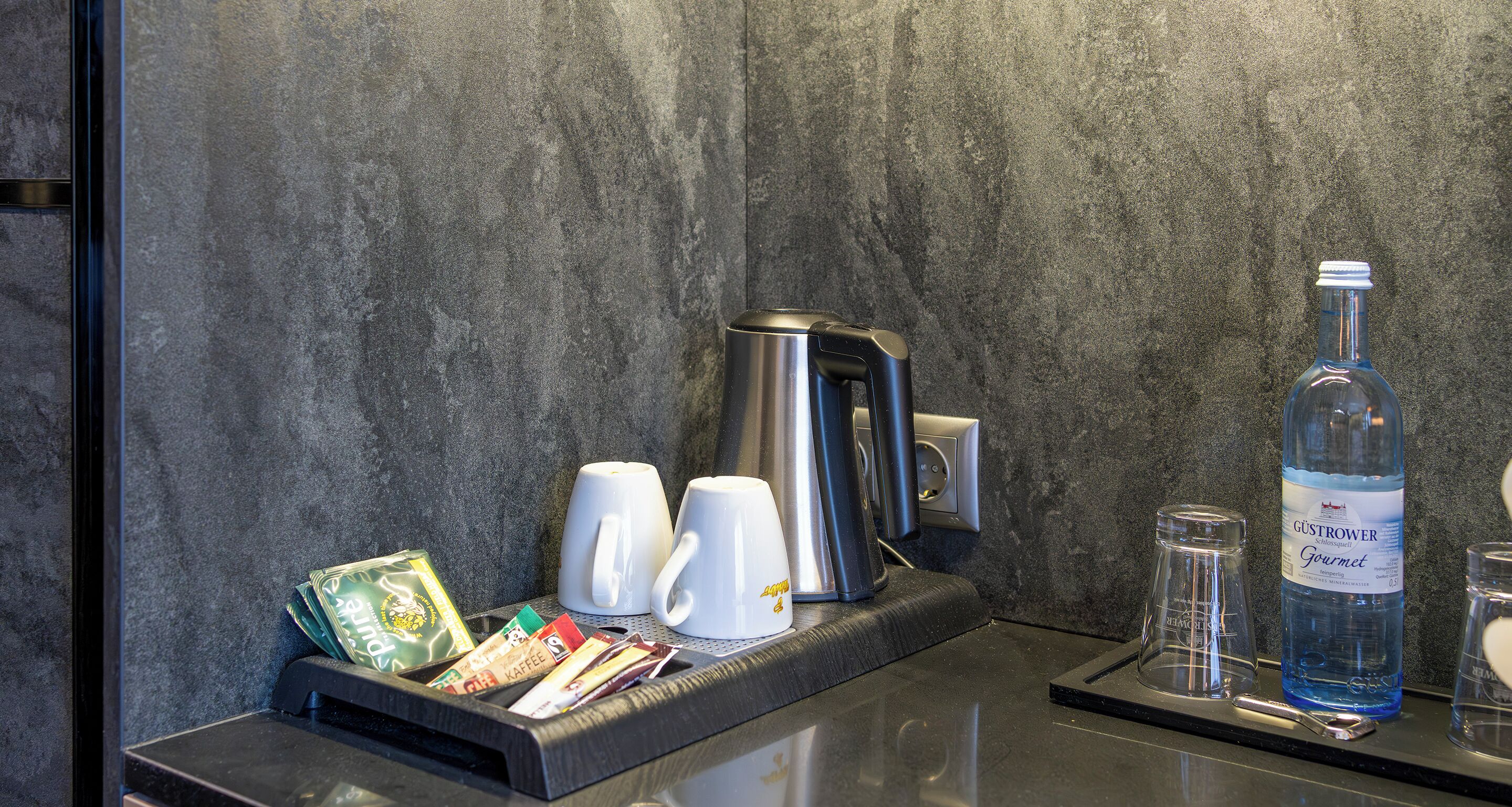 Ein Kaffee-Bereich mit zwei weißen Tassen, einer Kaffeemaschine und einer Wasserflasche auf einer Arbeitsplatte.