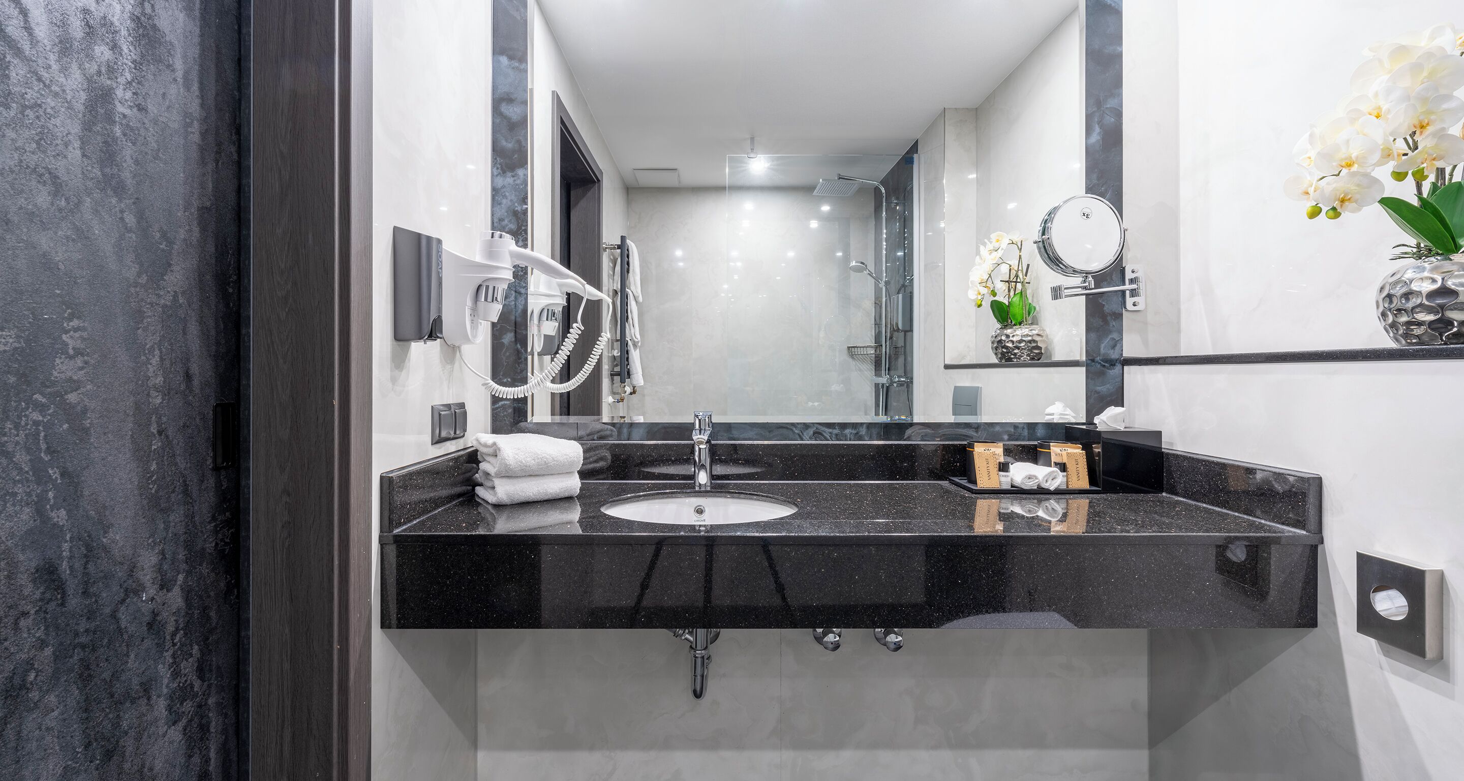 A modern bathroom with a black marble countertop, a sink, a mirror, a hairdryer, and a vase with white flowers.
