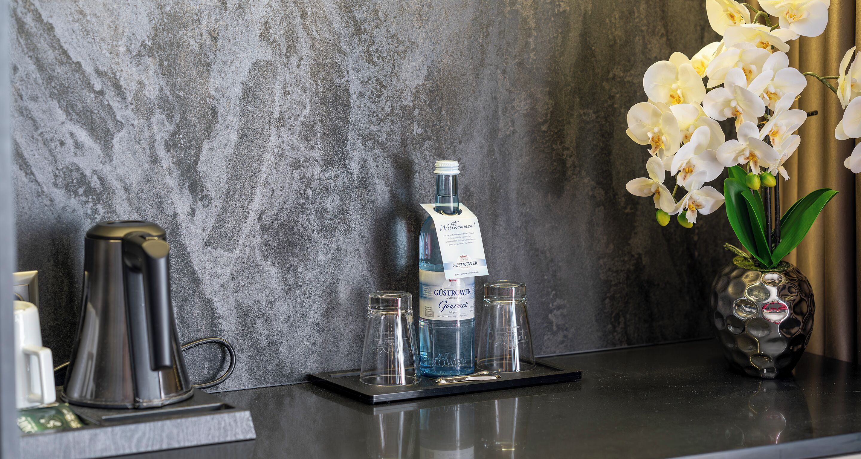 Un comptoir de cuisine avec une machine à café, une bouteille d’eau et un vase contenant des orchidées blanches.