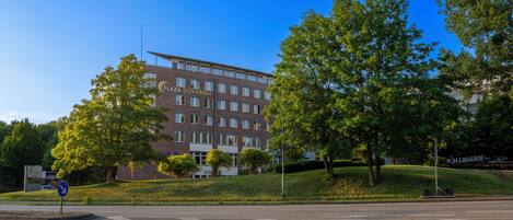 Een meerlaags hotel met een opvallend 'Plaza Schwerin' bord, omringd door weelderige begroeiing en een heldere blauwe lucht.
