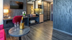 Une chambre d’hôtel moderne dotée d’une télévision à écran plat, d’un minibar, d’un fauteuil en velours rouge, d’une table ronde avec une bouteille et des verres, et d’un plancher en bois.
