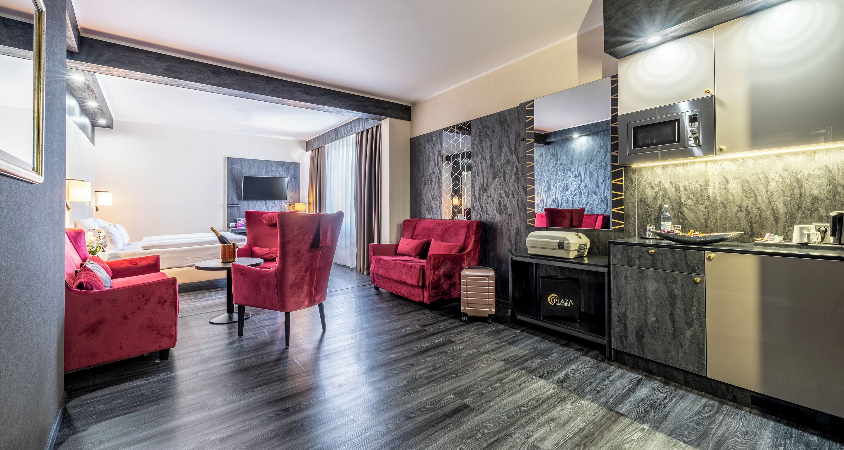 Una habitación de hotel moderna con piso de madera oscura, sillas tapizadas en rojo, un televisor de pantalla plana en la pared, un minibar y una cocina integrada.