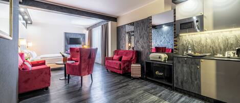 Una habitación de hotel moderna con piso de madera oscura, sillas tapizadas en rojo, un televisor de pantalla plana en la pared, un minibar y una cocina integrada.