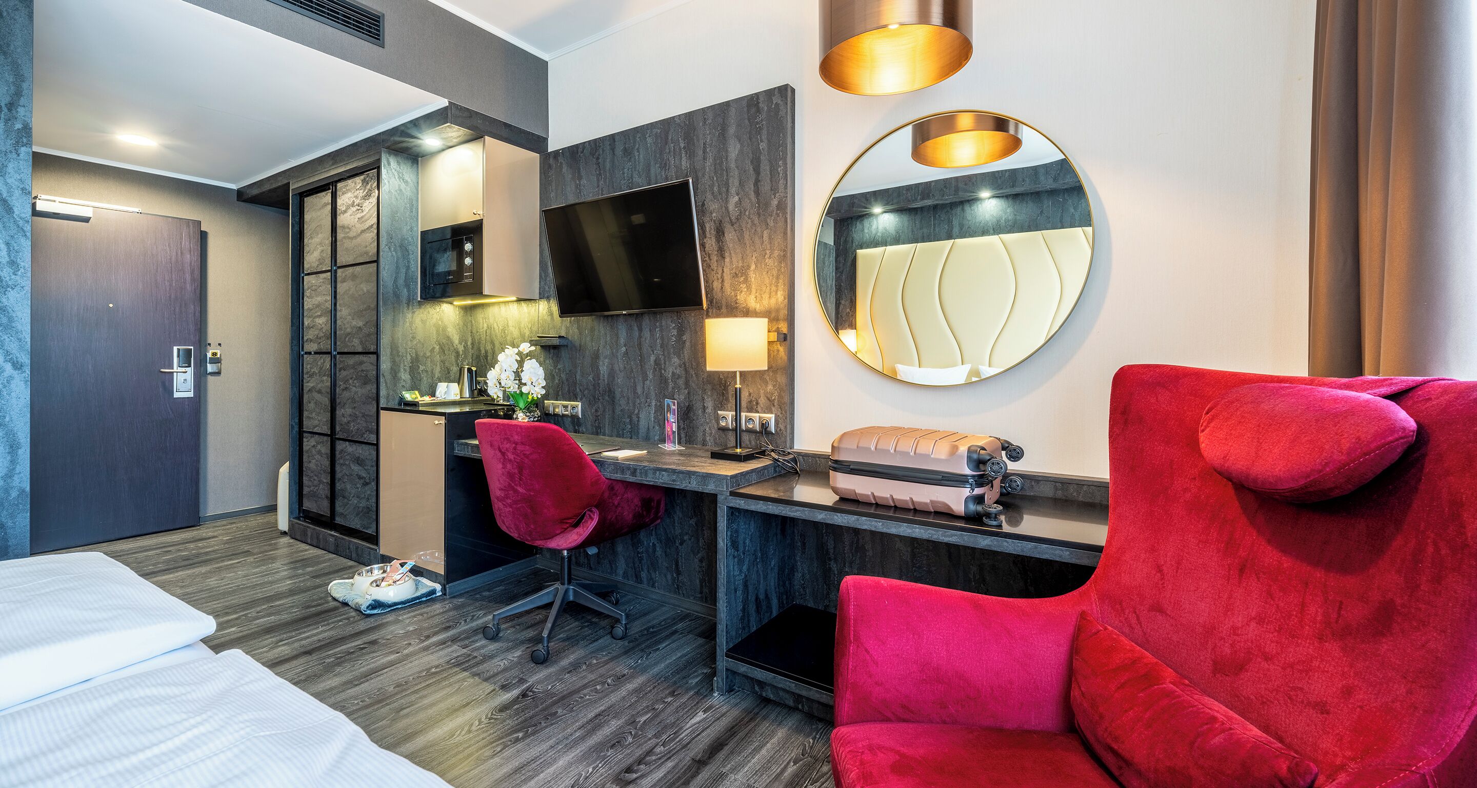 Une chambre d’hôtel moderne avec un lit, un bureau, une chaise rouge, une télévision, un miroir et une valise posée sur le bureau.