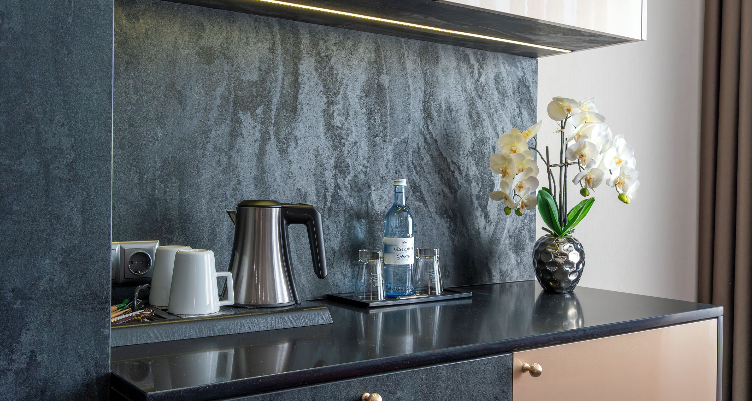 Une cuisine moderne avec un dosseret en marbre, une machine à café, une bouilloire, un vase orné d’orchidées et une bouteille d’eau.