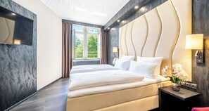 A modern hotel room with a large bed, a flat-screen TV, a nightstand with a lamp, and a view of greenery outside. - PLAZA Premium Schwerin (Schwerin)