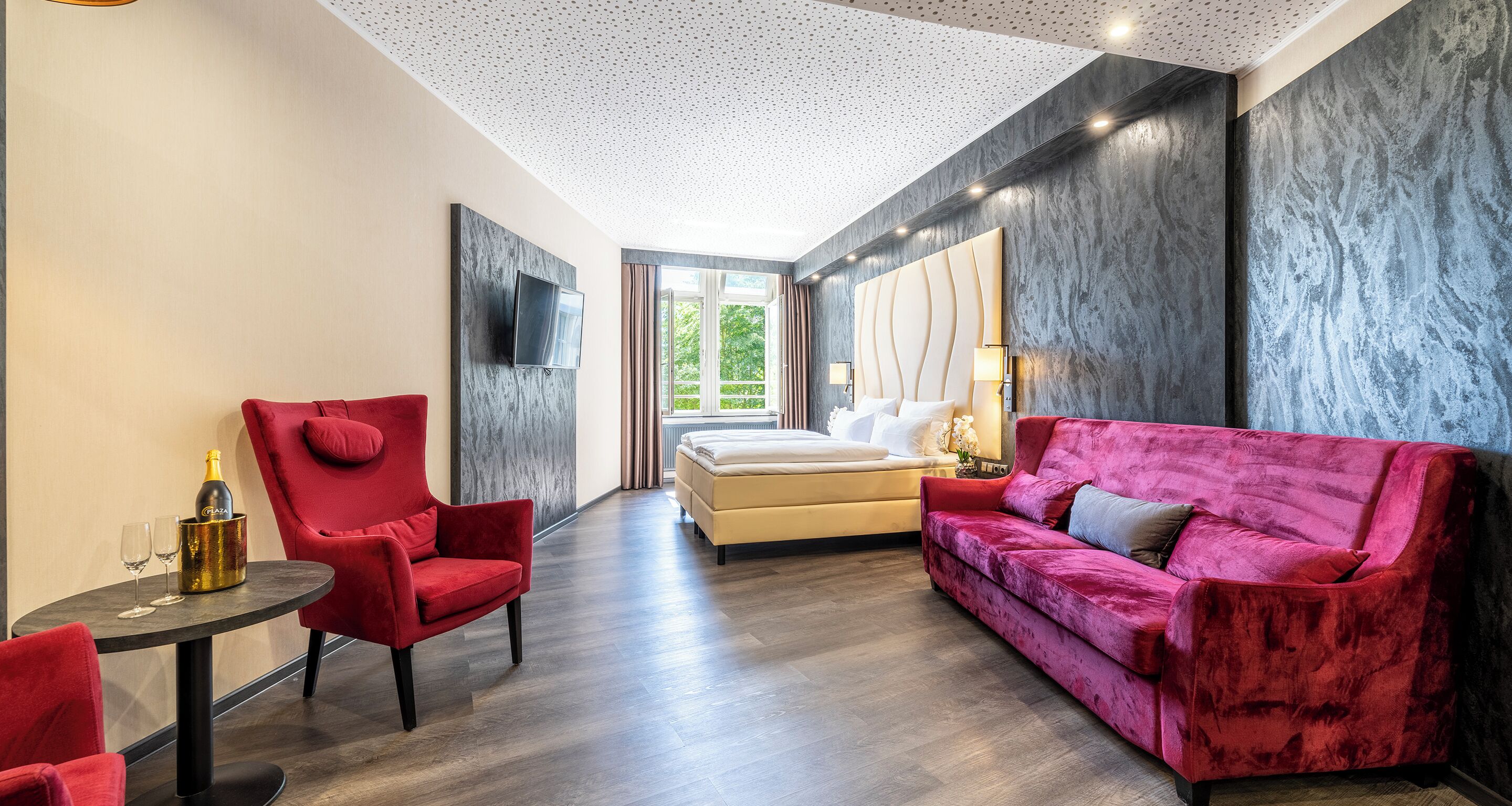 Une chambre d’hôtel moderne avec un lit, un fauteuil rouge, un canapé rose, une petite table avec une bouteille et des verres, et un téléviseur fixé au mur.