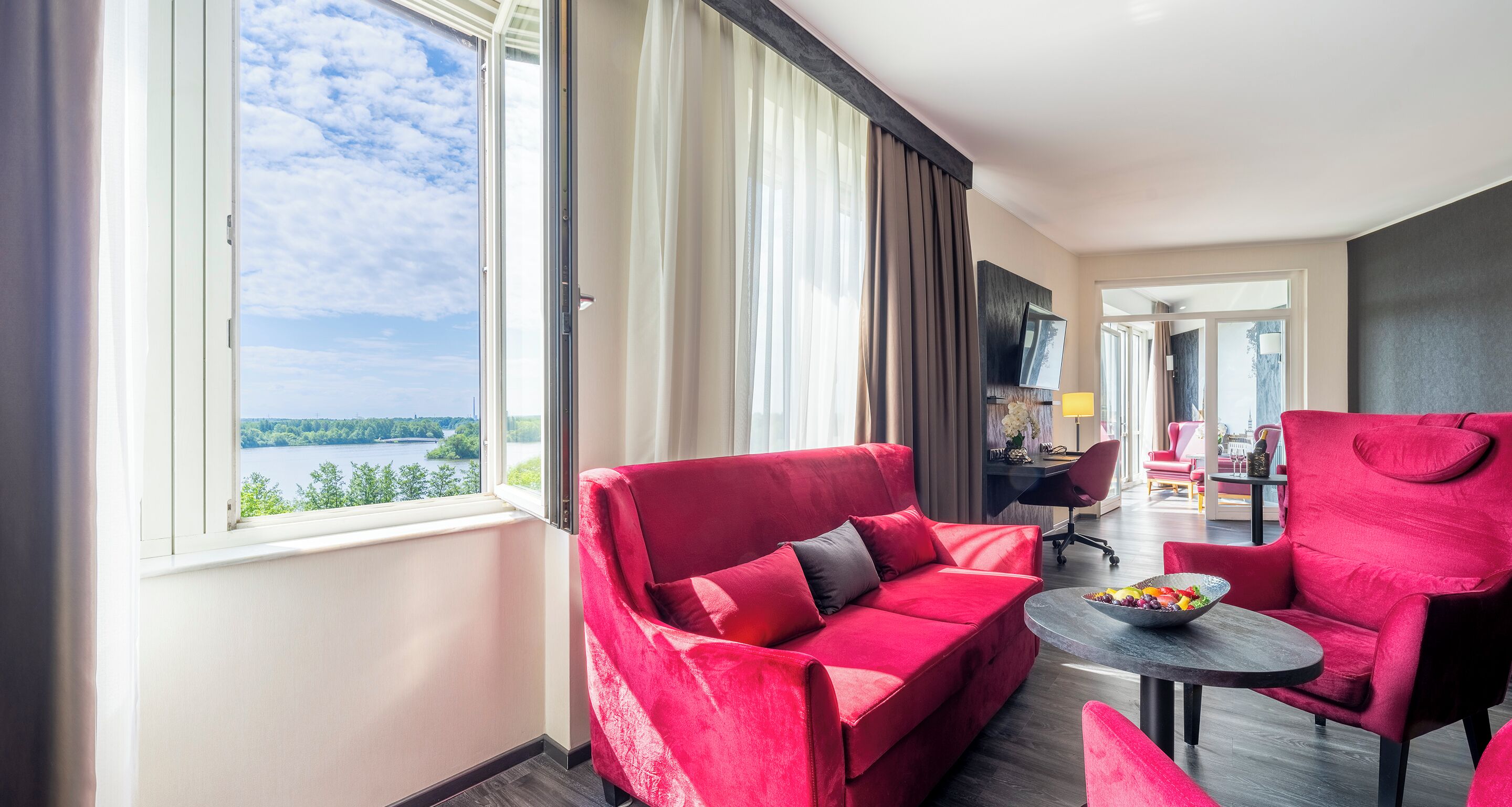 A hotel room with a red sofa, a small round table with a bowl of fruit, and a view of a lake through a large window.