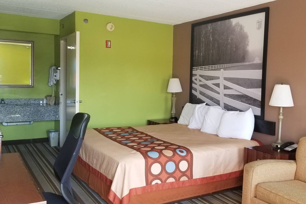 A hotel room with a bed, a chair, a sofa, a desk, a mirror, a hairdryer, and a painting on the wall.