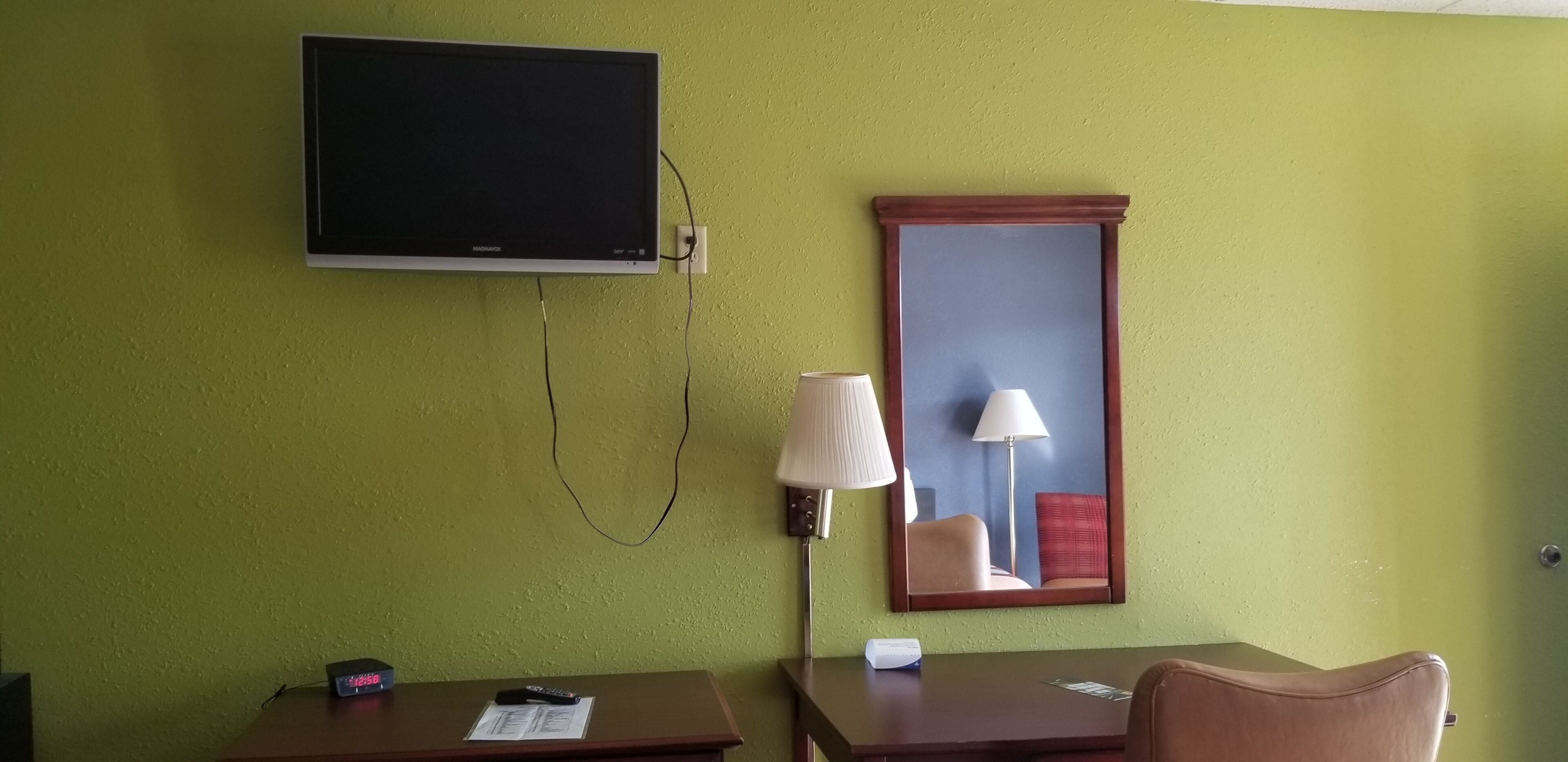 Une chambre d’hôtel équipée d’un téléviseur à écran plat fixé au mur, d’une table de chevet avec une lampe, d’une chaise et d’une petite table sur laquelle se trouvent une télécommande et une boîte de mouchoirs.