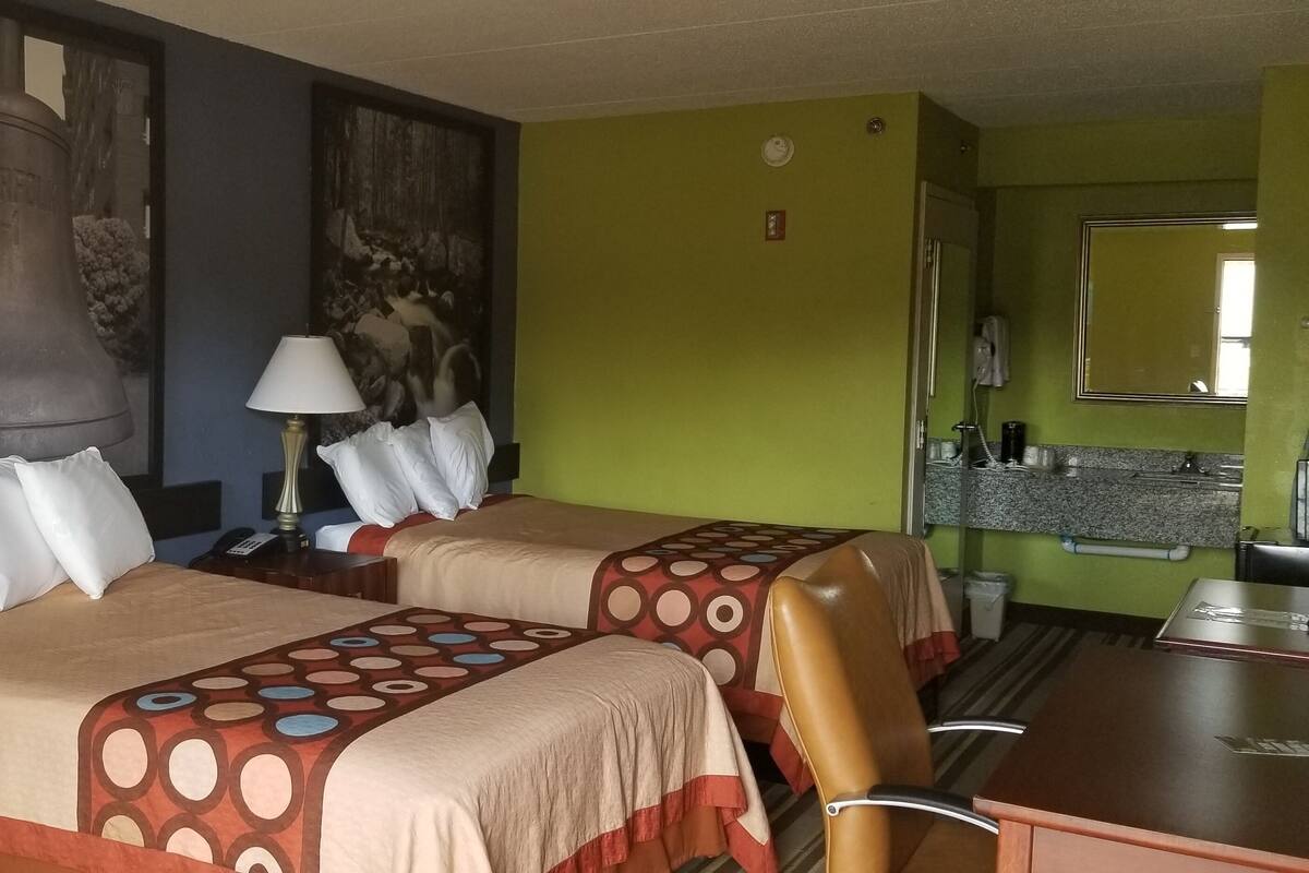 A hotel room with two beds, a desk, and a chair. There are two nightstands with lamps and a telephone. The walls are painted green, and there is a large painting of a bell.