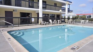 Piscina com espreguiçadeiras e guarda-sóis, um edifício com vários andares e varandas, e um céu claro.