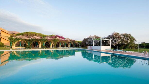 A swimming pool with clear water, surrounded by covered seating areas and lush greenery. - Resort Marina di Castello Golf & SPA (Castel Volturno)