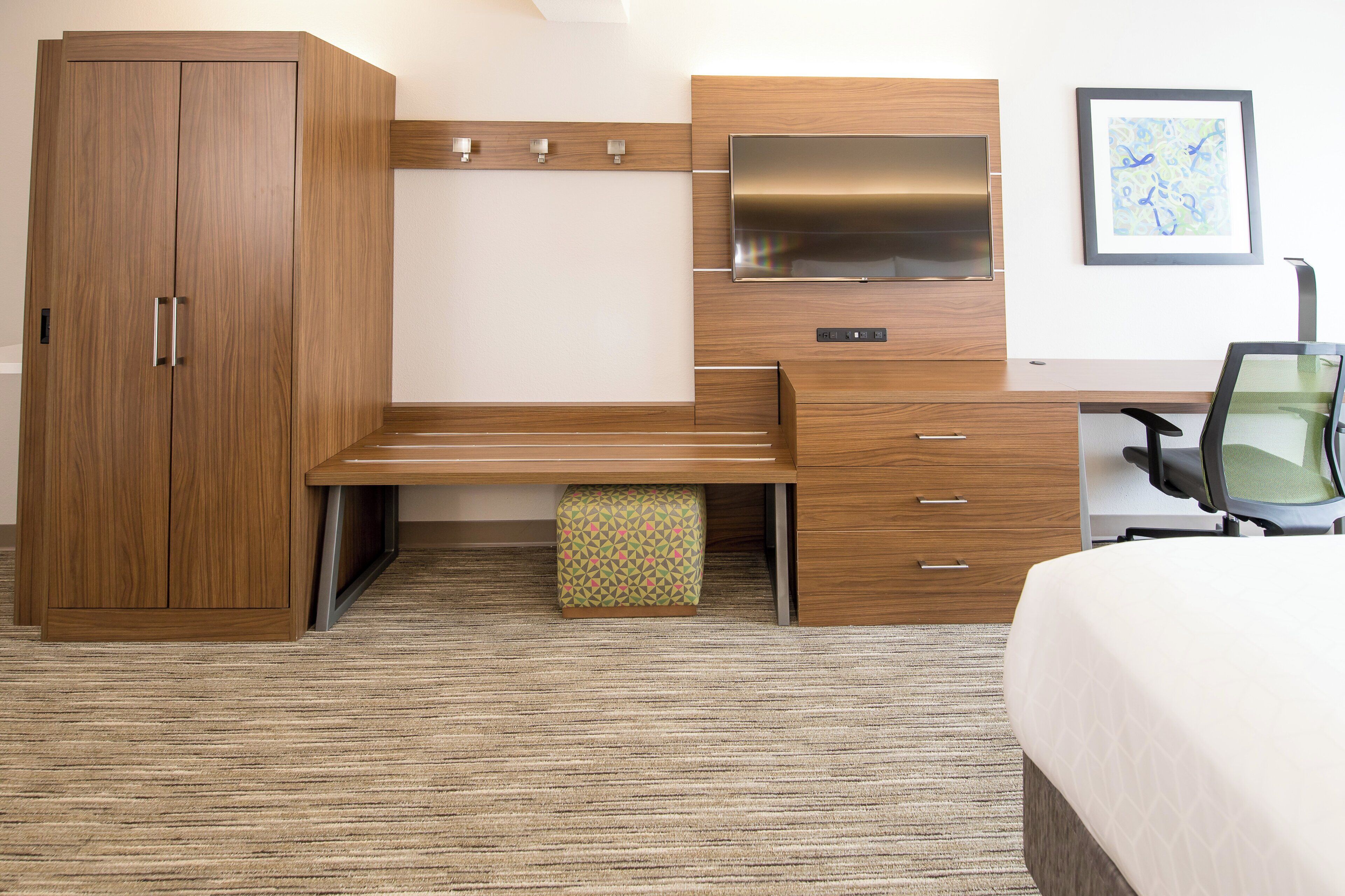 Une chambre d’hôtel comprenant une armoire en bois, un bureau avec des tiroirs, un banc, un petit pouf, une chaise et un lit.